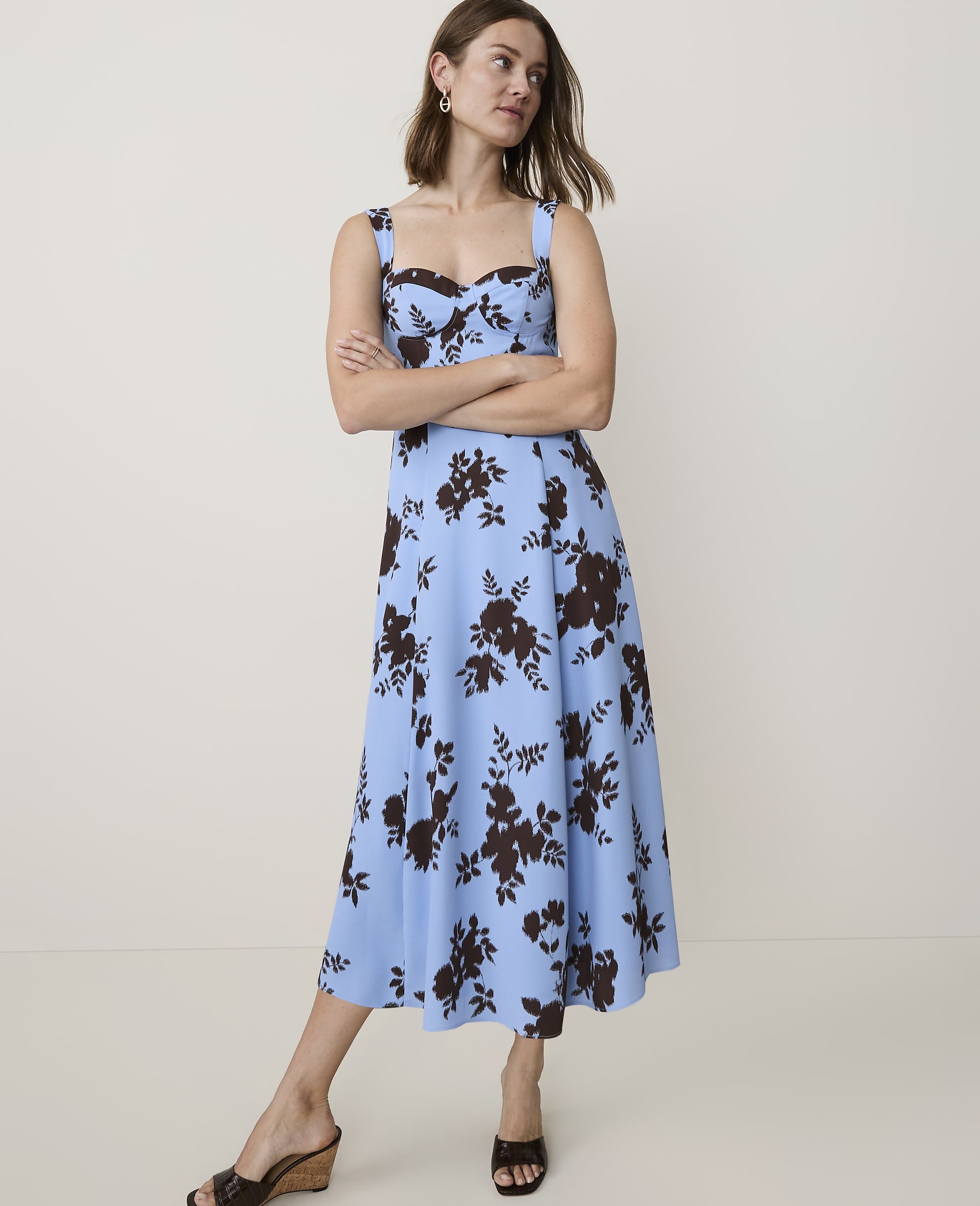 A woman in a blue floral dress stands with her arms crossed against a plain, light-colored background.