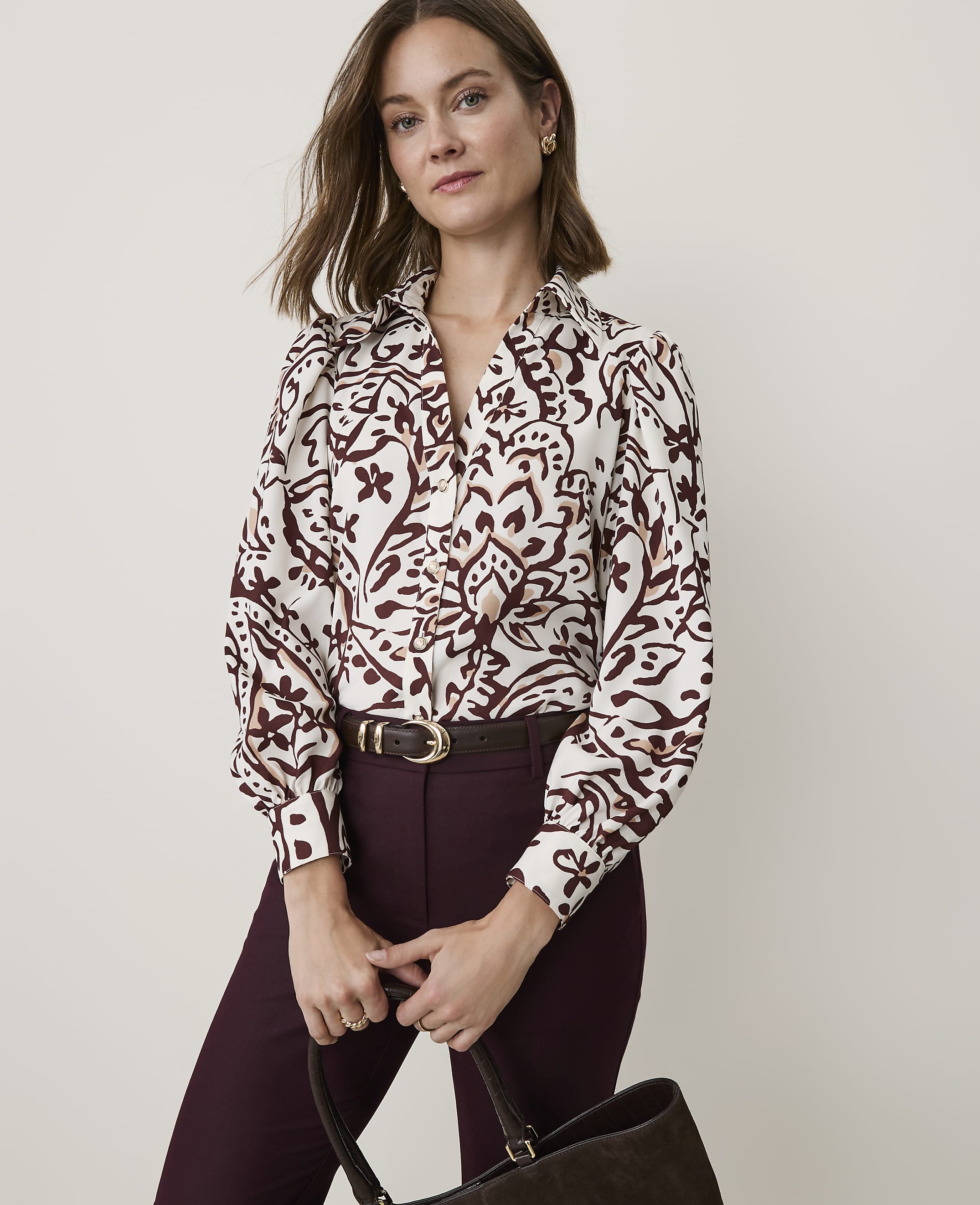 A woman in a patterned blouse and burgundy pants holds a dark brown bag in front of a plain, light-colored background.