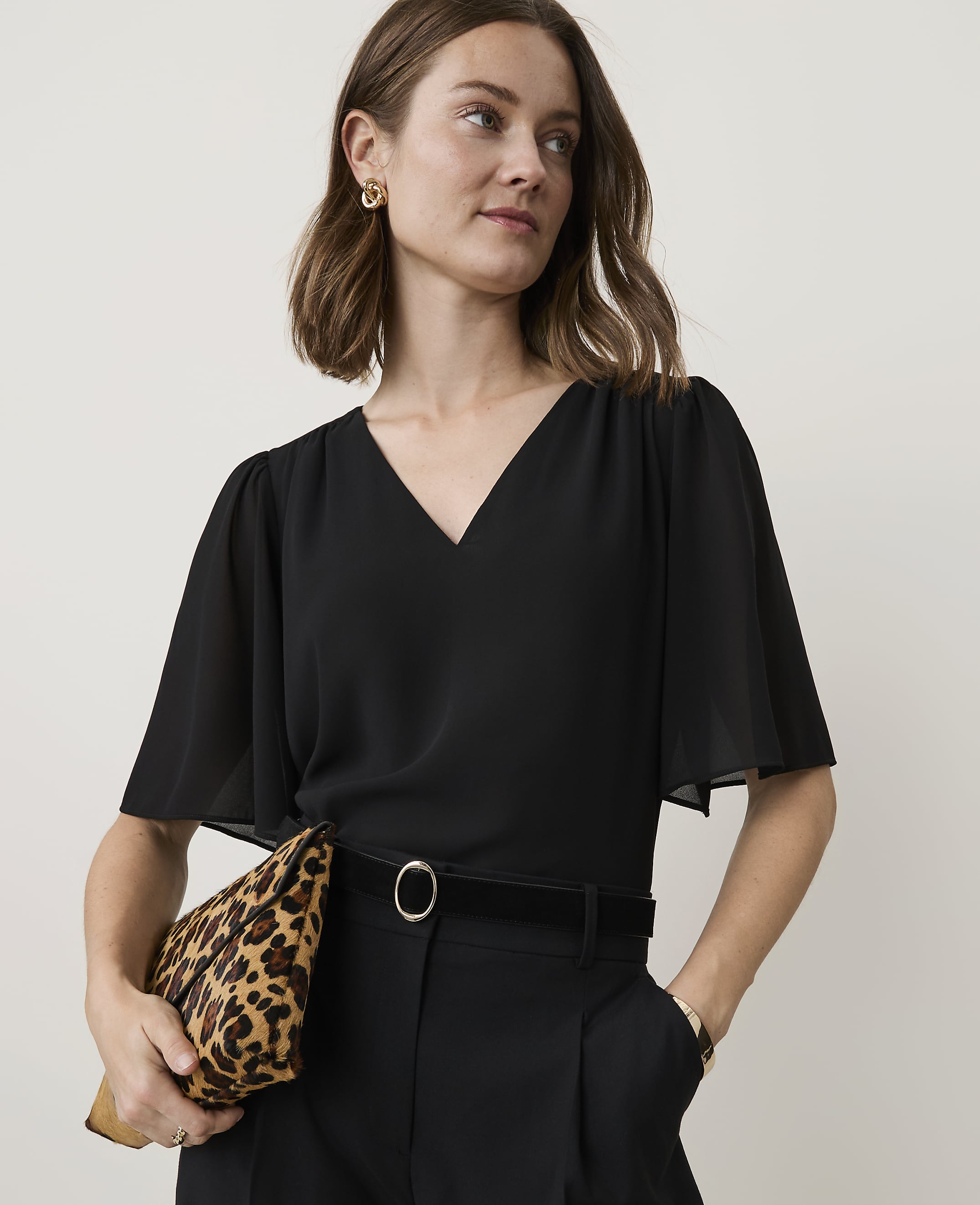 A woman in a black v-neck top and black pants holds a leopard print clutch in front of a plain background.