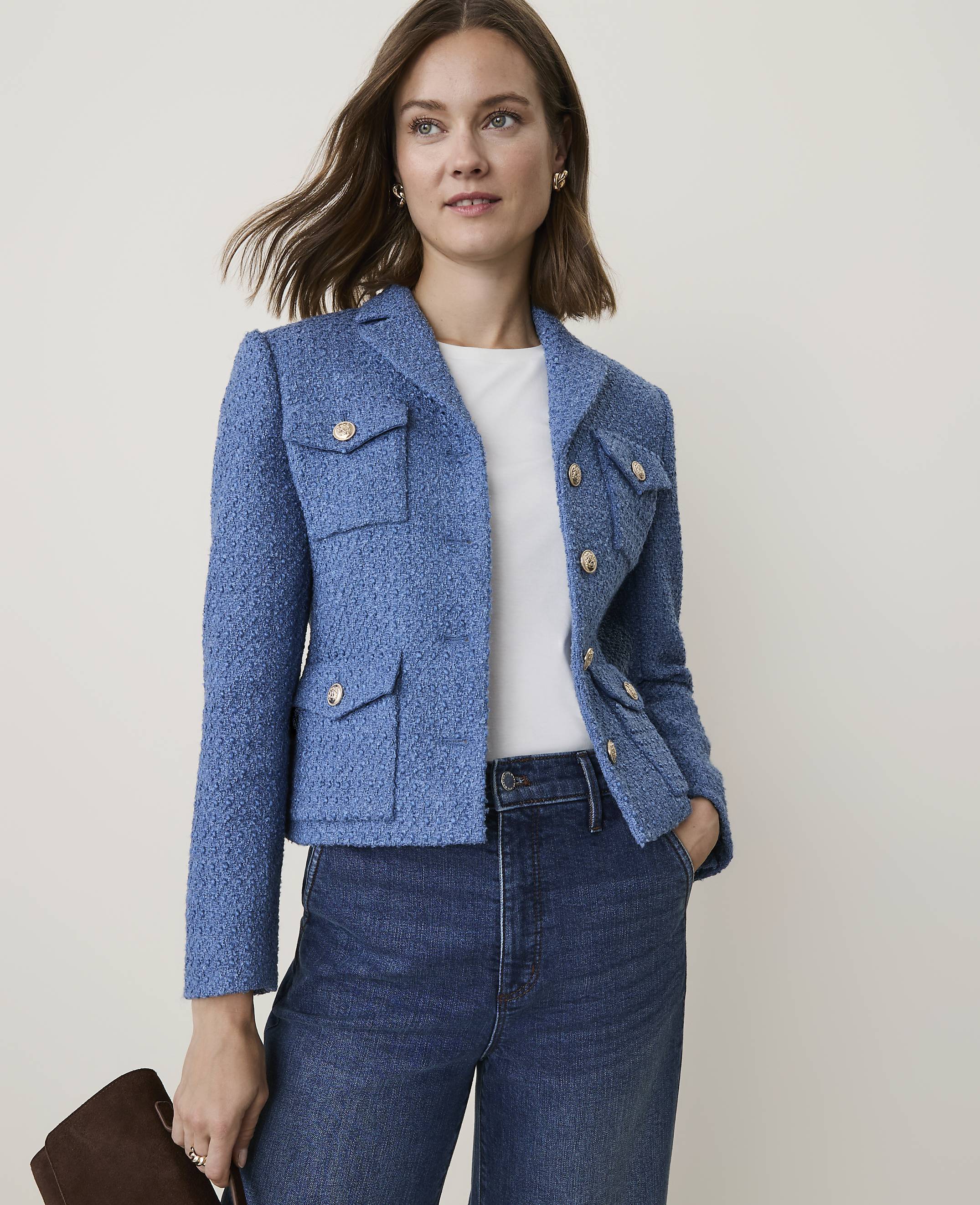 A woman in a blue tweed jacket and jeans stands against a plain, light background, holding a brown clutch.