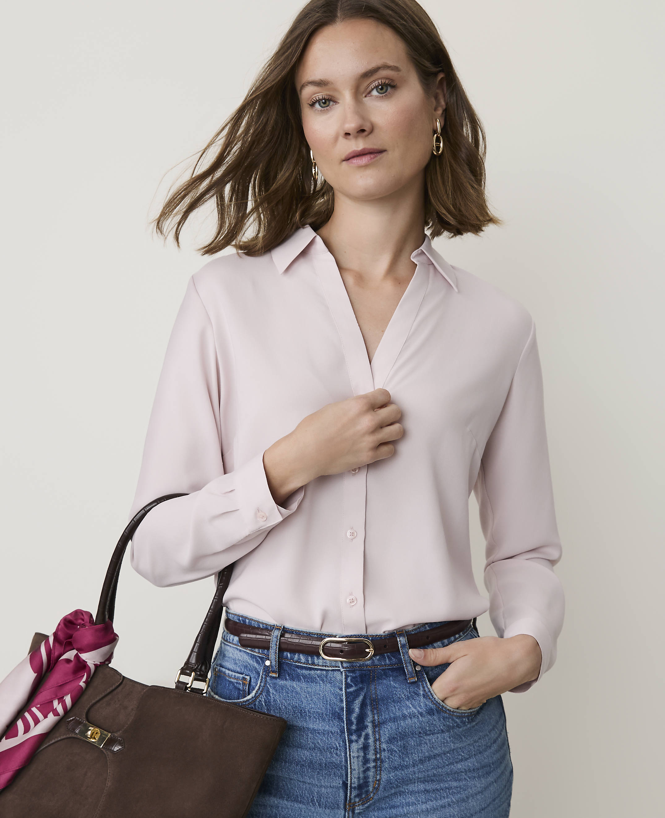 A woman in a light pink blouse and jeans stands in front of a plain, light-colored background, with a brown handbag on her left side.