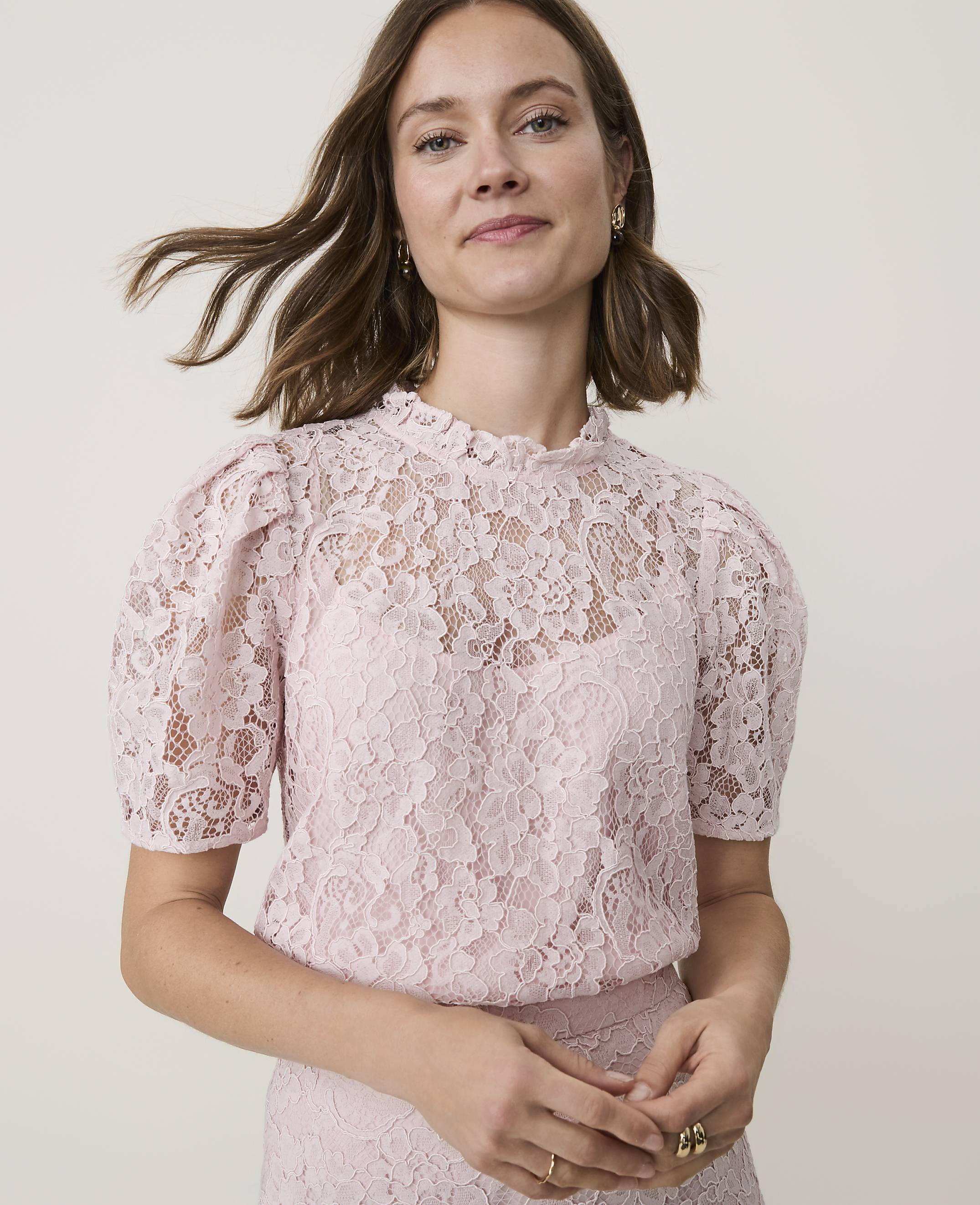 A woman in a pink lace top with puffed sleeves stands against a plain, light background.