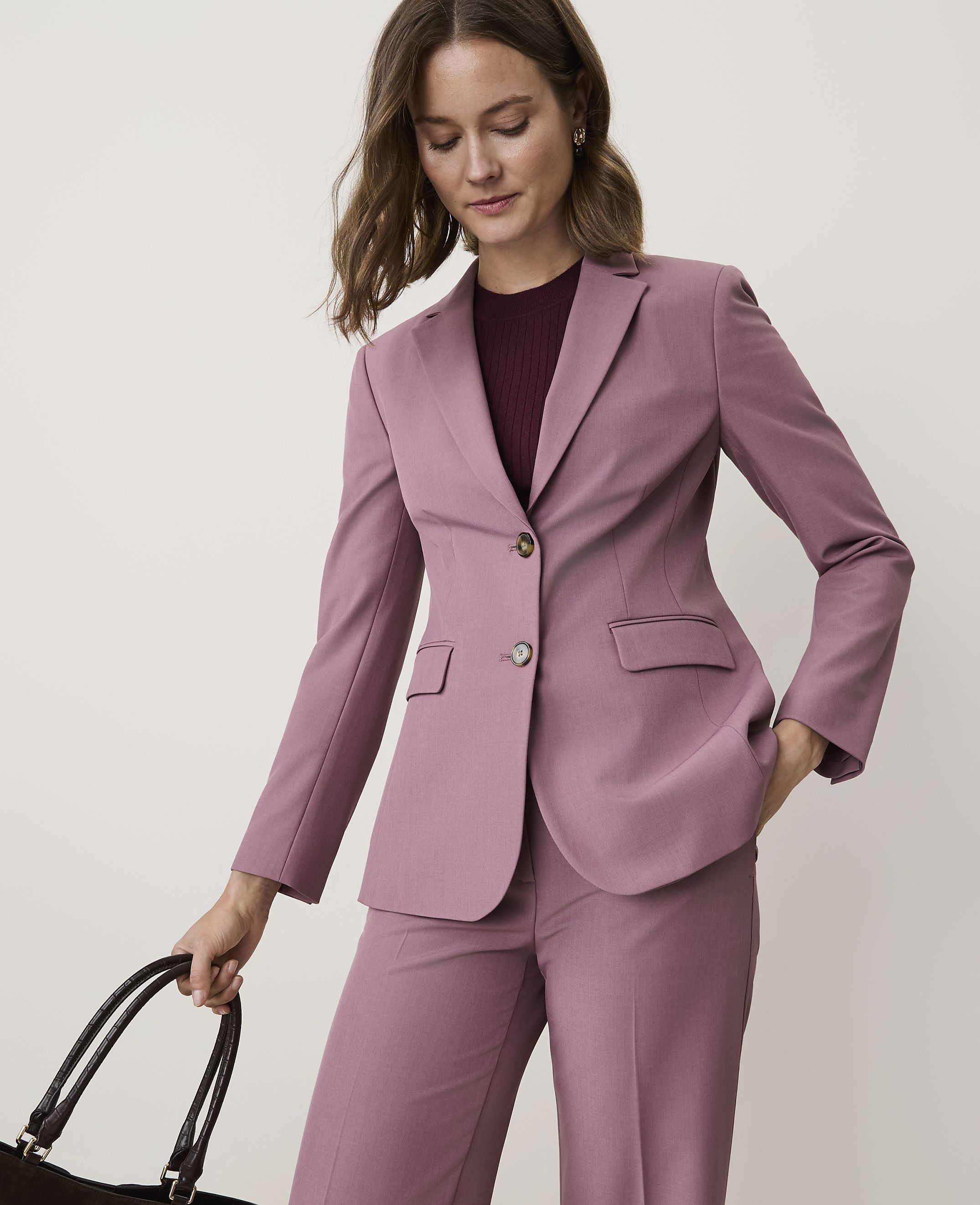 A woman in a mauve pantsuit with her hand in her pocket stands against a plain, light background, holding a dark tote bag.