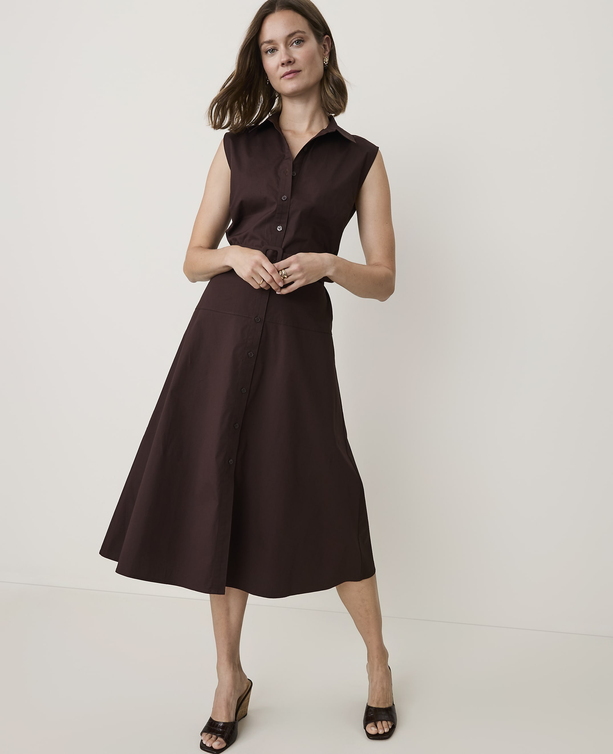 A woman in a brown sleeveless shirt dress stands against a plain white background.