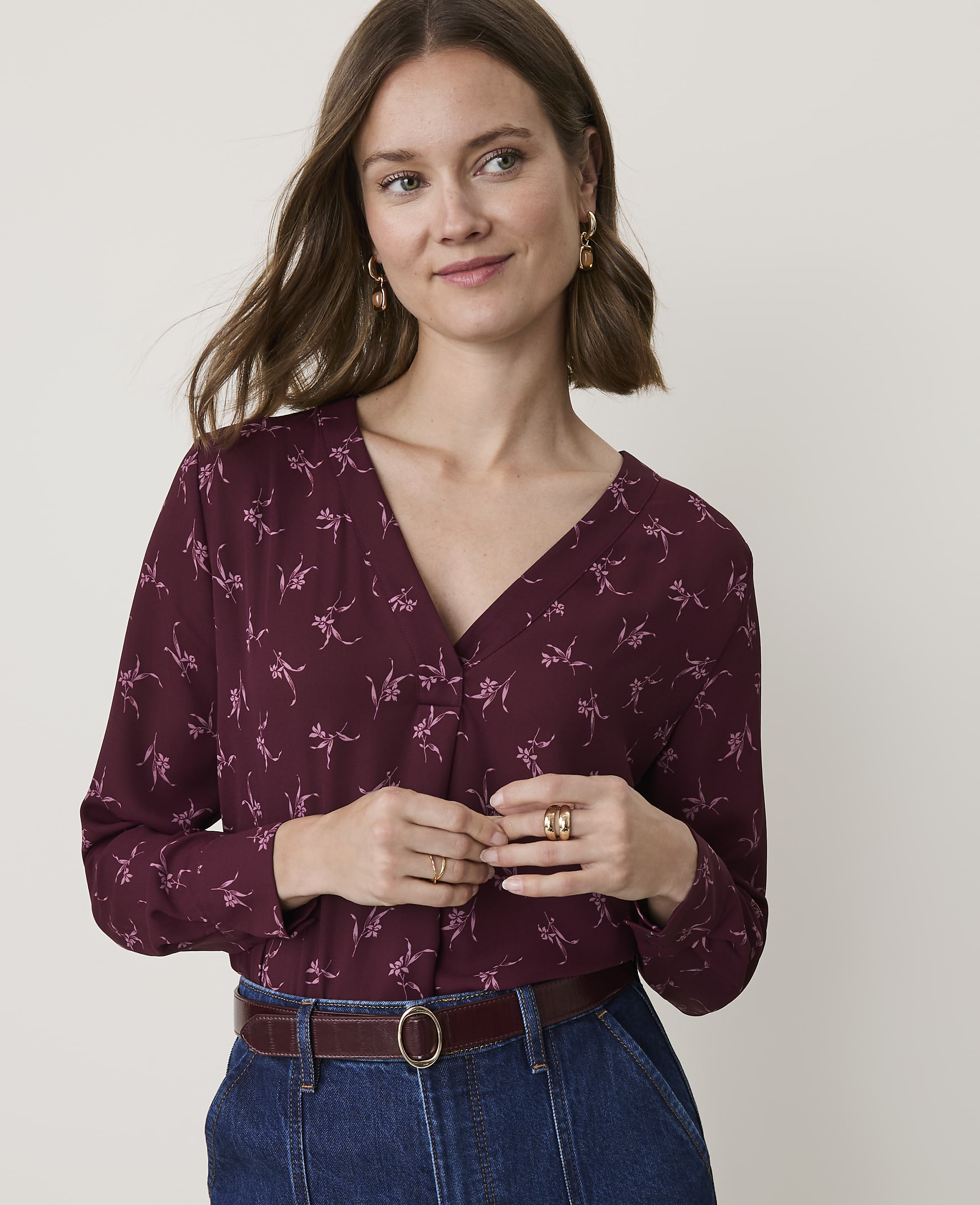 A woman in a burgundy floral blouse and denim jeans stands against a plain, light background.