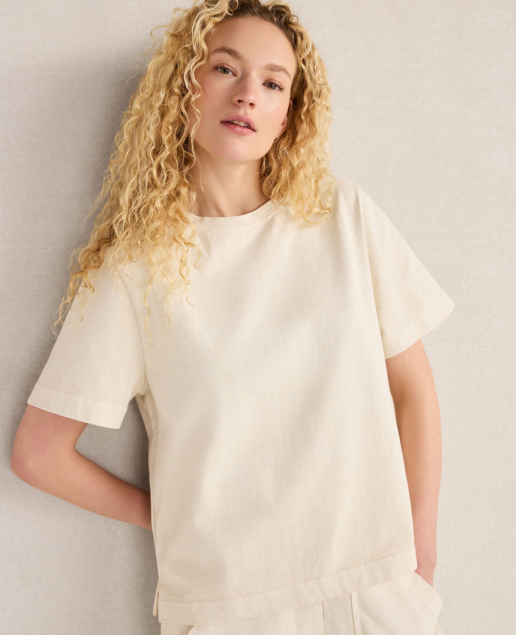 A young woman with curly blonde hair wears a cream-colored t-shirt and pants, leaning against a textured gray wall.