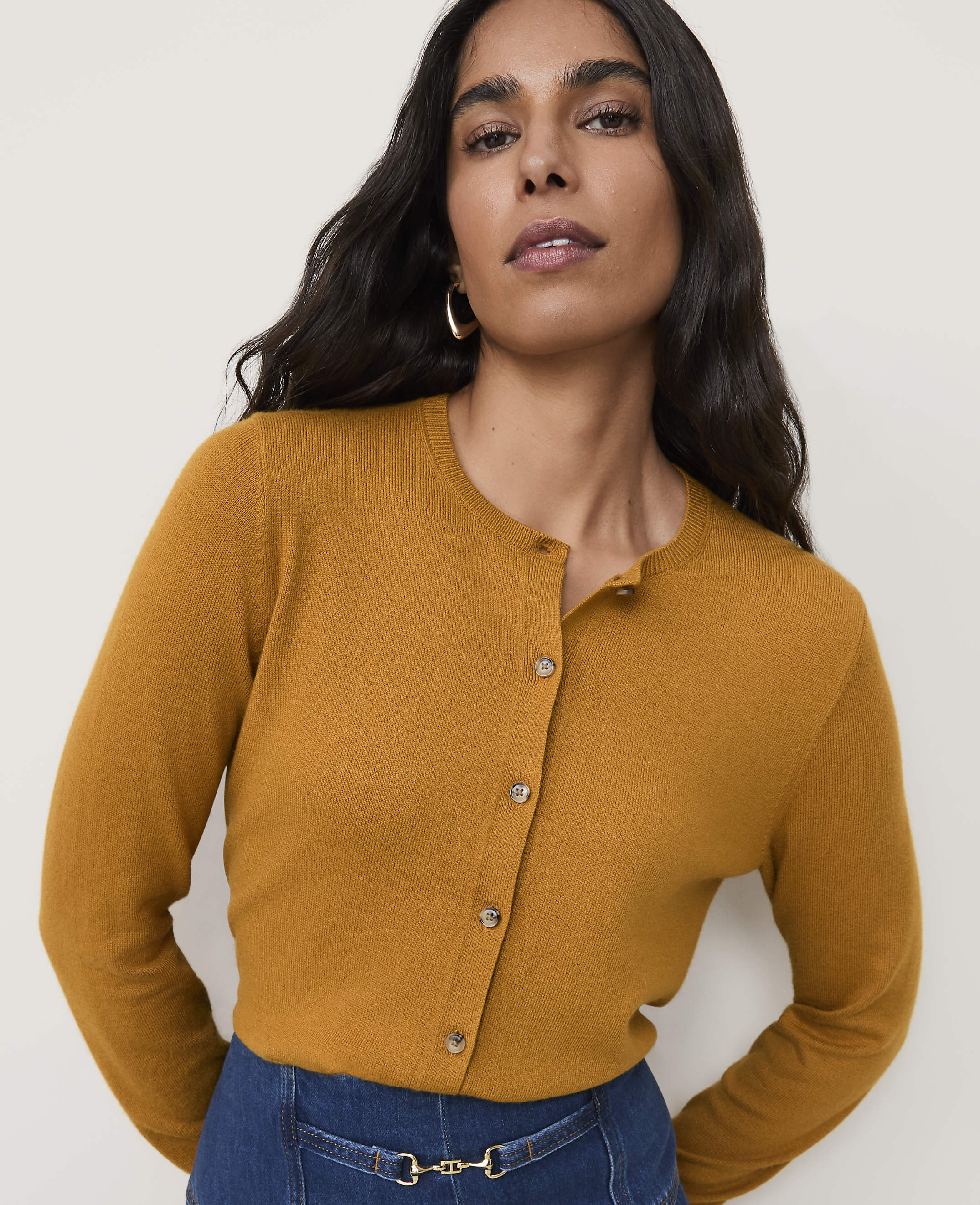 A woman in a mustard-colored cardigan stands against a plain, light background.