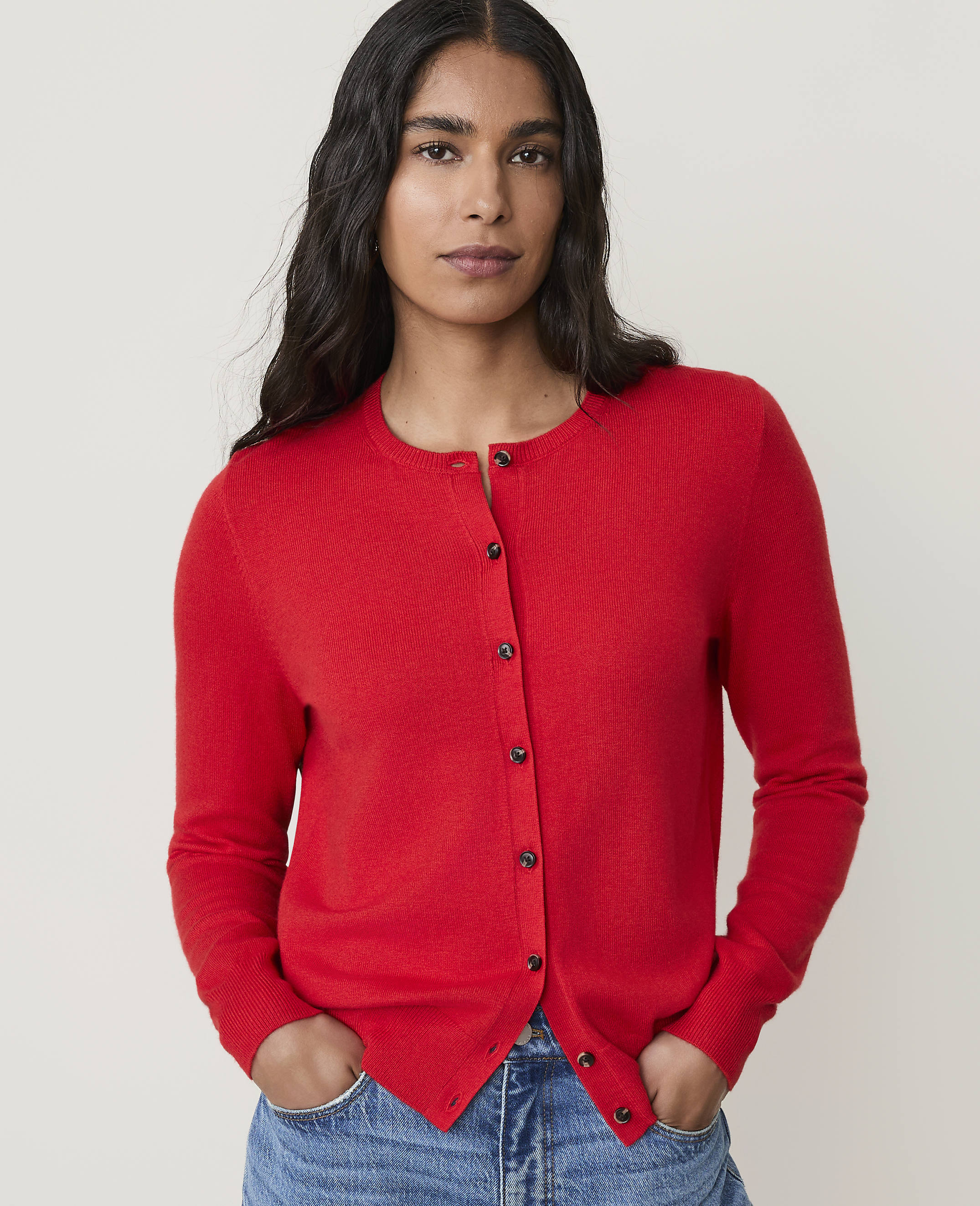 A woman in a red cardigan and blue jeans stands against a plain white background.