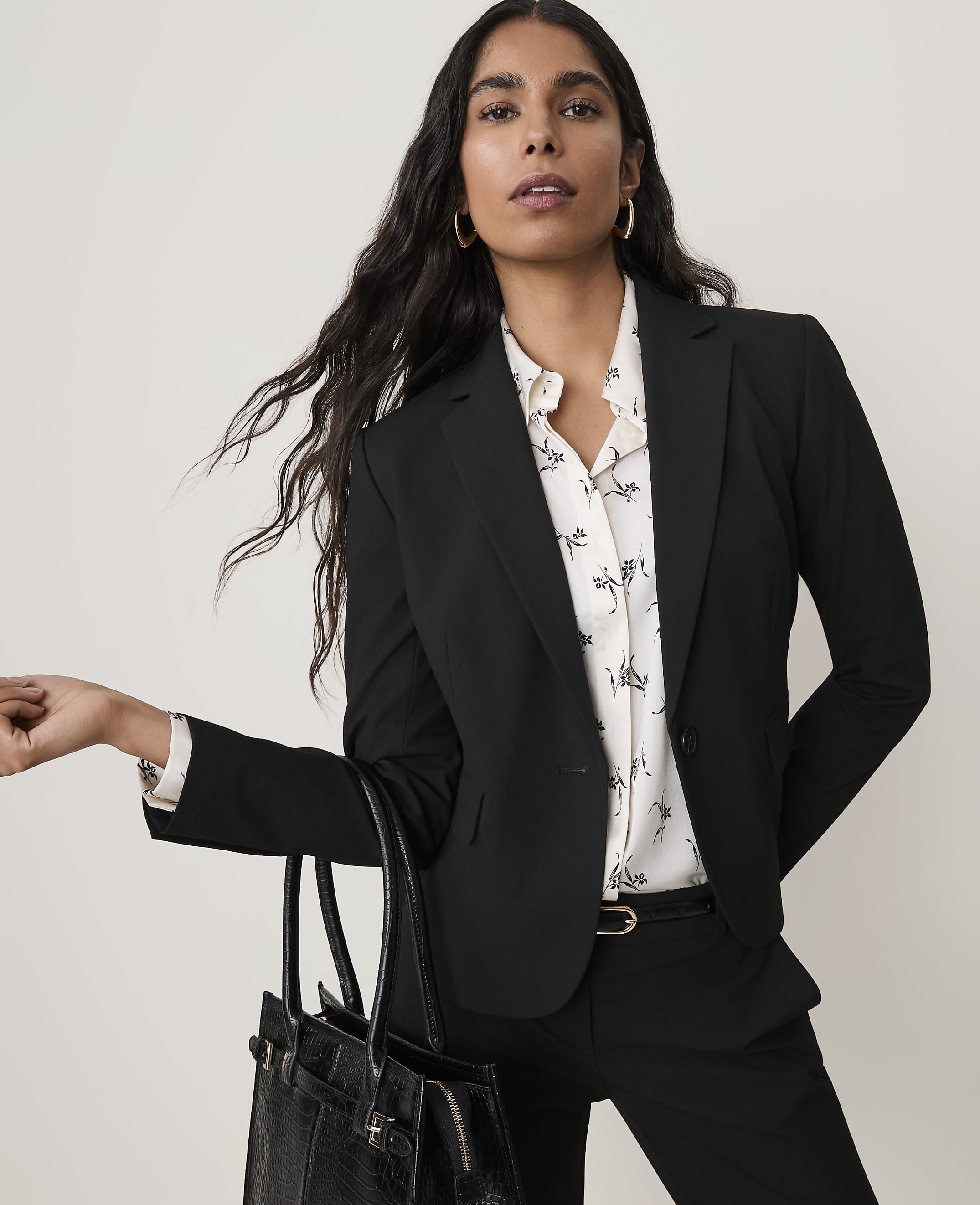 A woman in a black blazer and patterned blouse holds a black handbag in front of a plain white background.