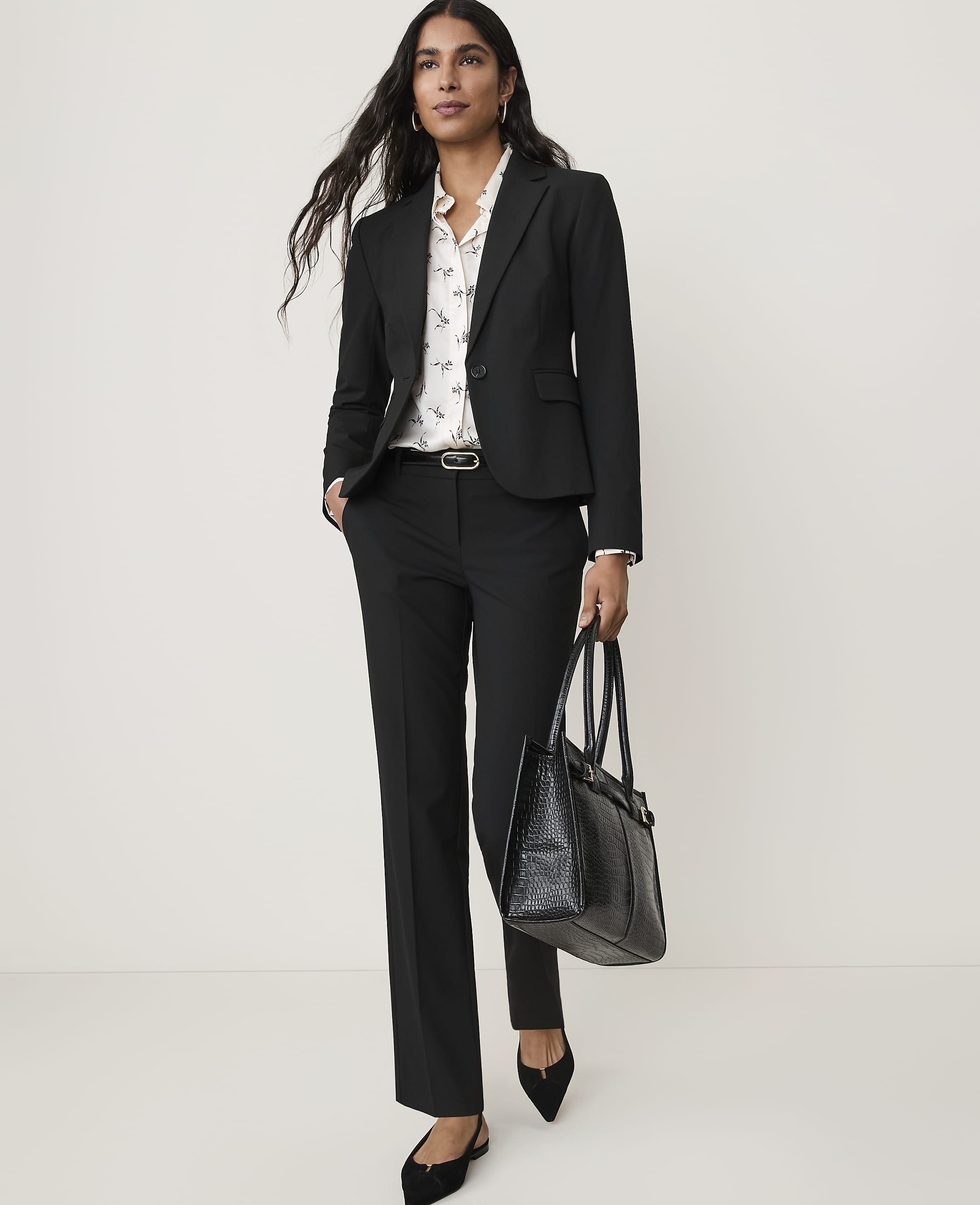 A woman in a black suit and patterned blouse stands with a black tote bag against a plain white background.