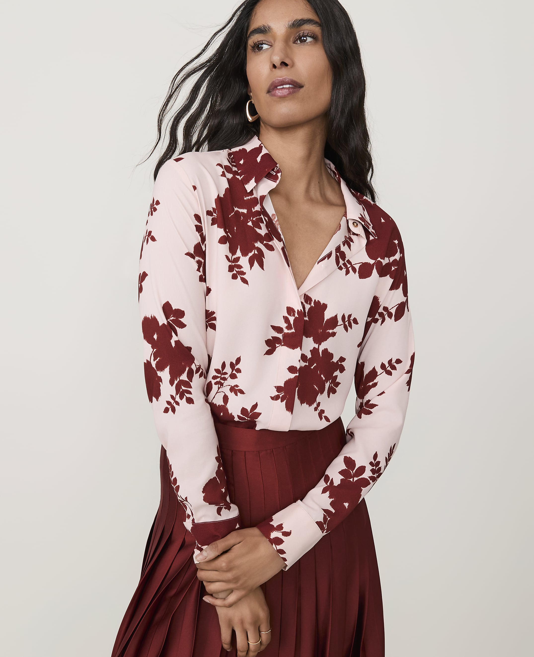 A woman in a floral blouse and pleated skirt stands against a plain background.