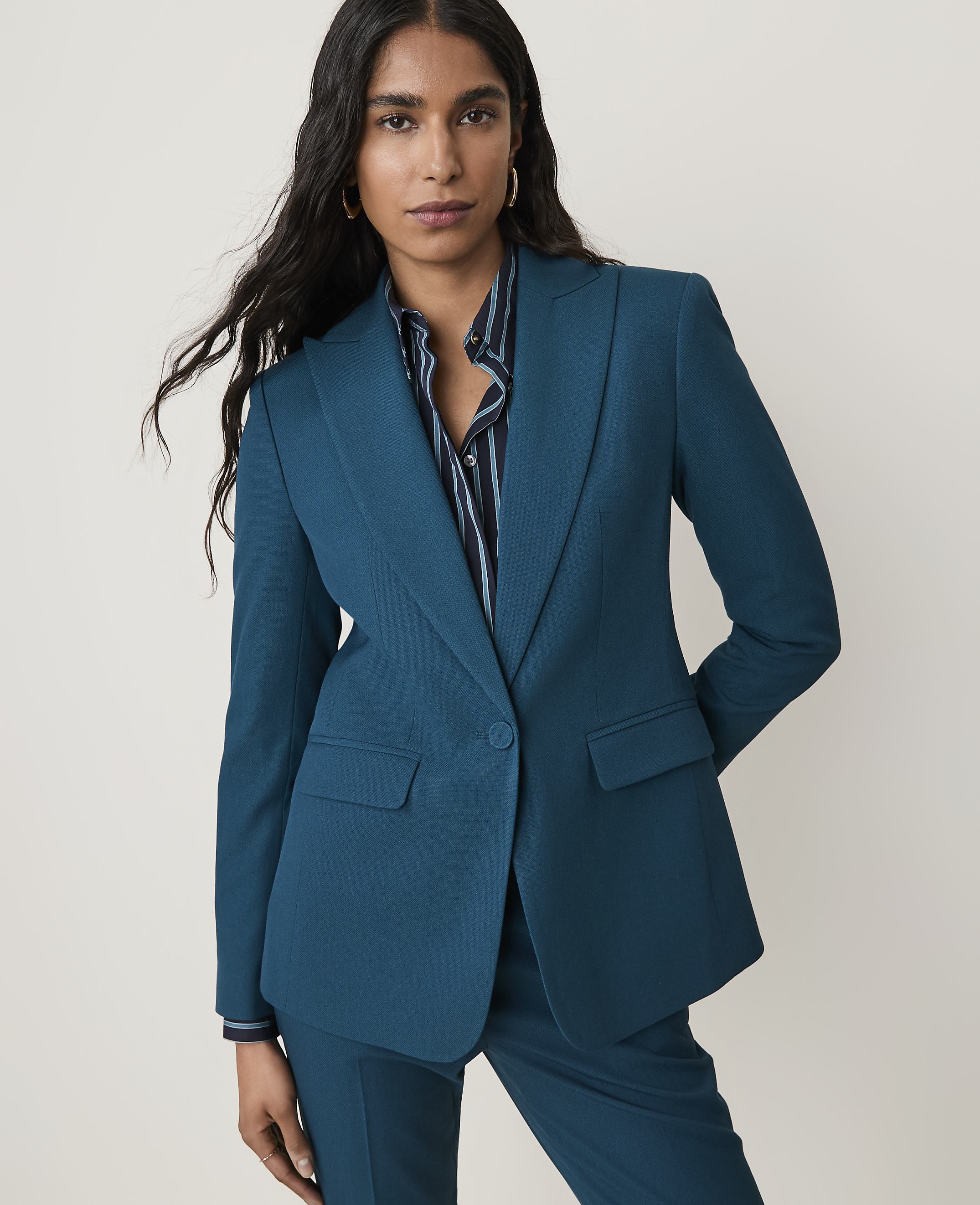 A woman in a teal blazer and striped shirt stands against a plain white background.