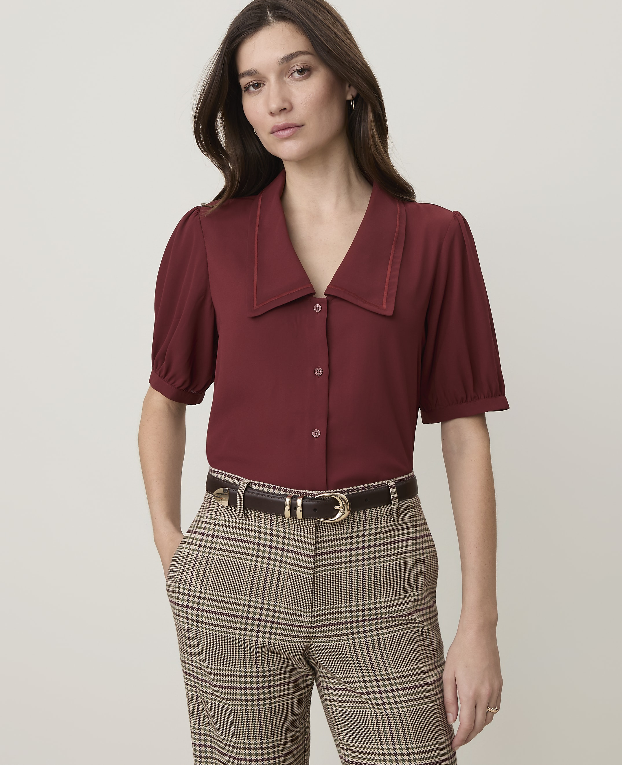 A woman in a maroon blouse and plaid pants stands against a plain, light background.
