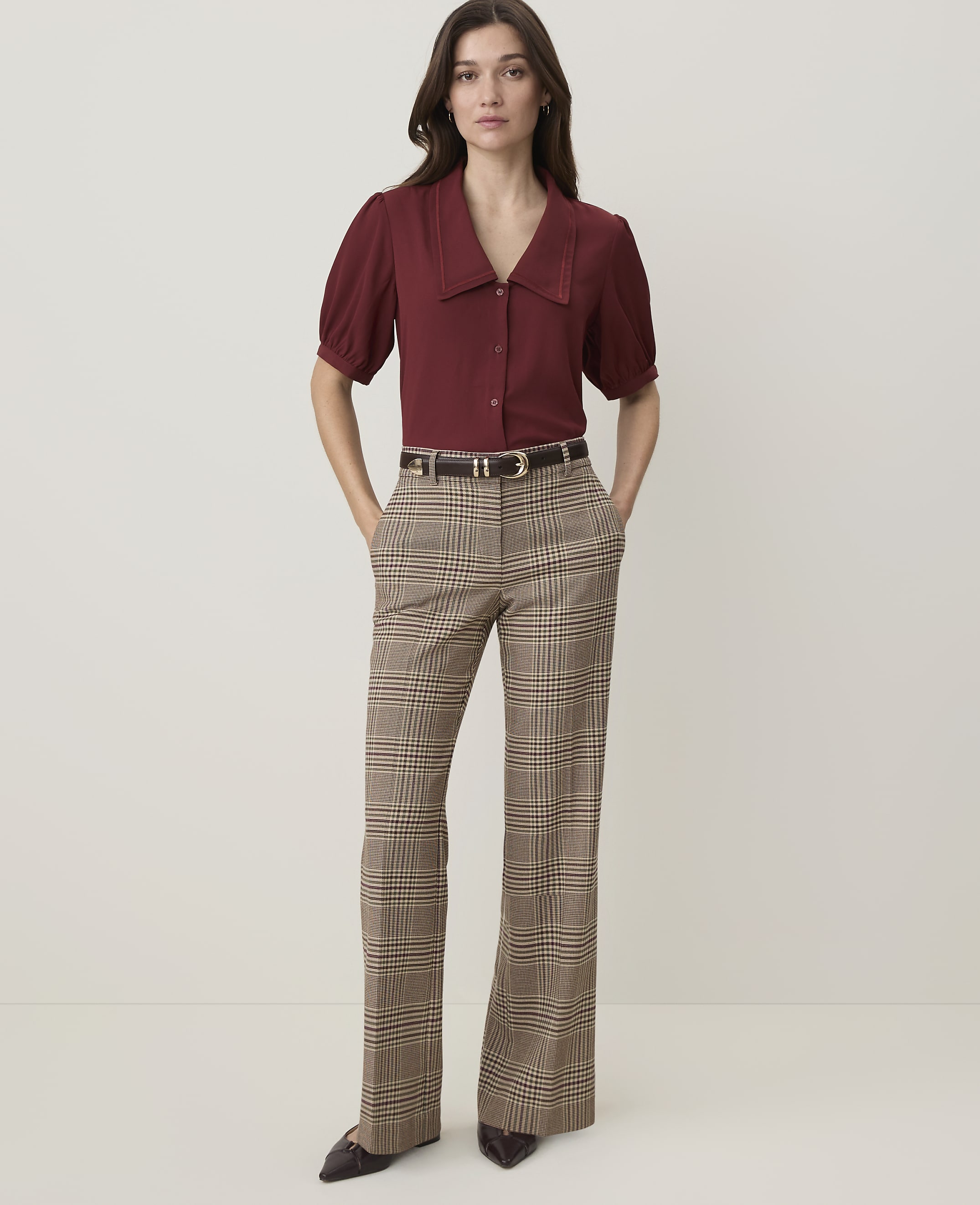 A woman in a maroon blouse and plaid pants stands against a plain, light-colored background.
