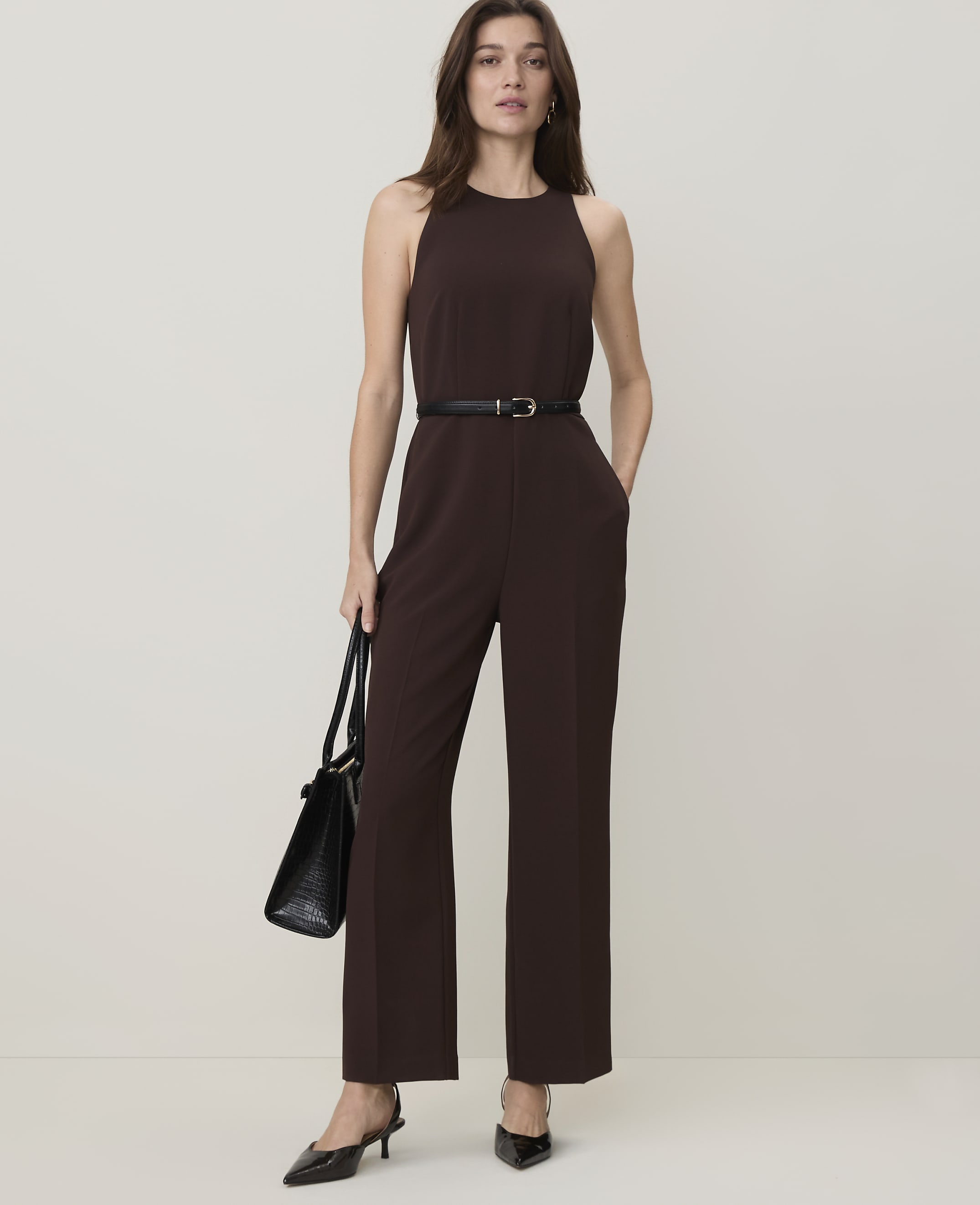 A woman in a brown jumpsuit and black heels stands with a black handbag against a plain, light brown background.