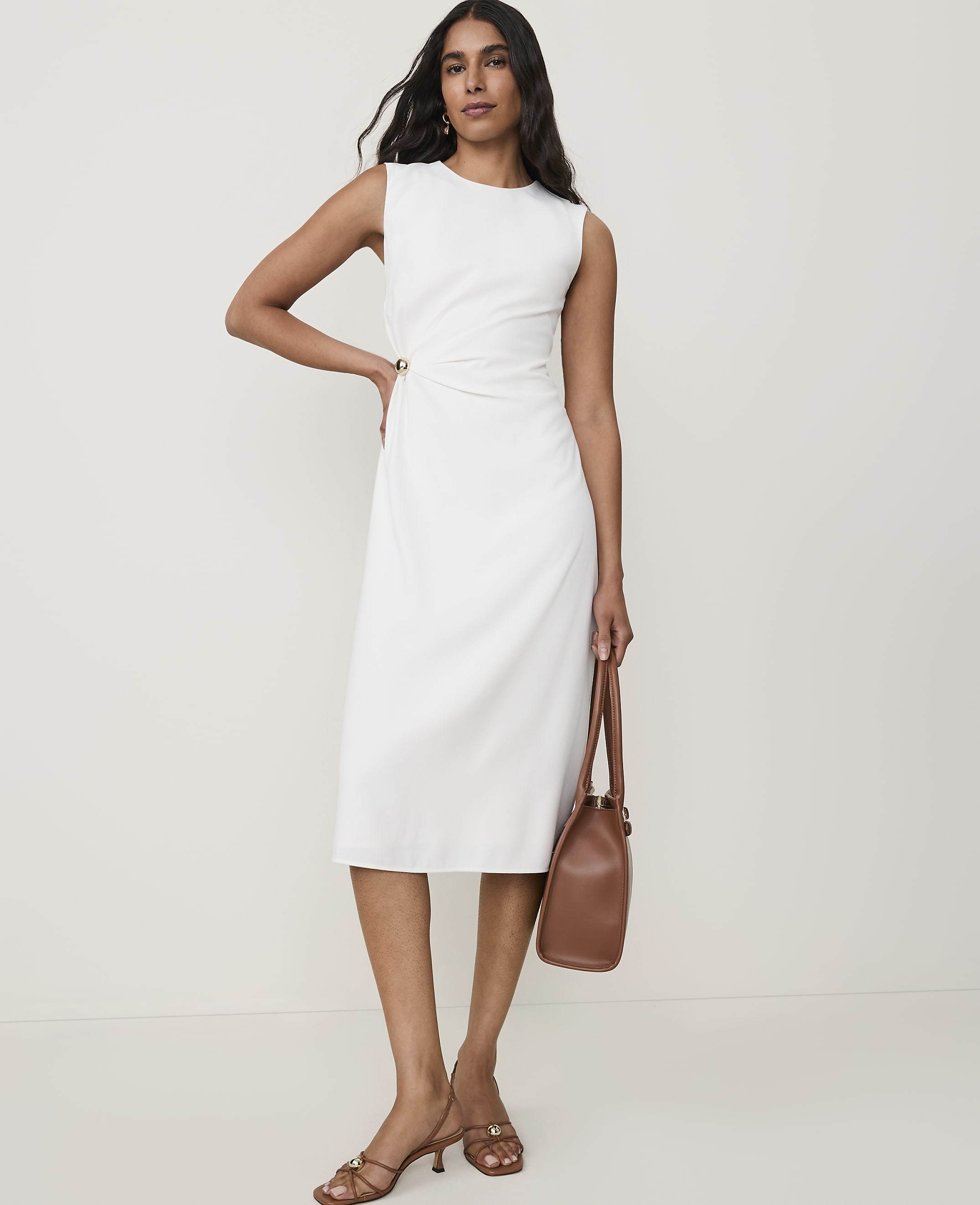 A woman in a white dress and brown sandals stands with a brown purse against a plain background.