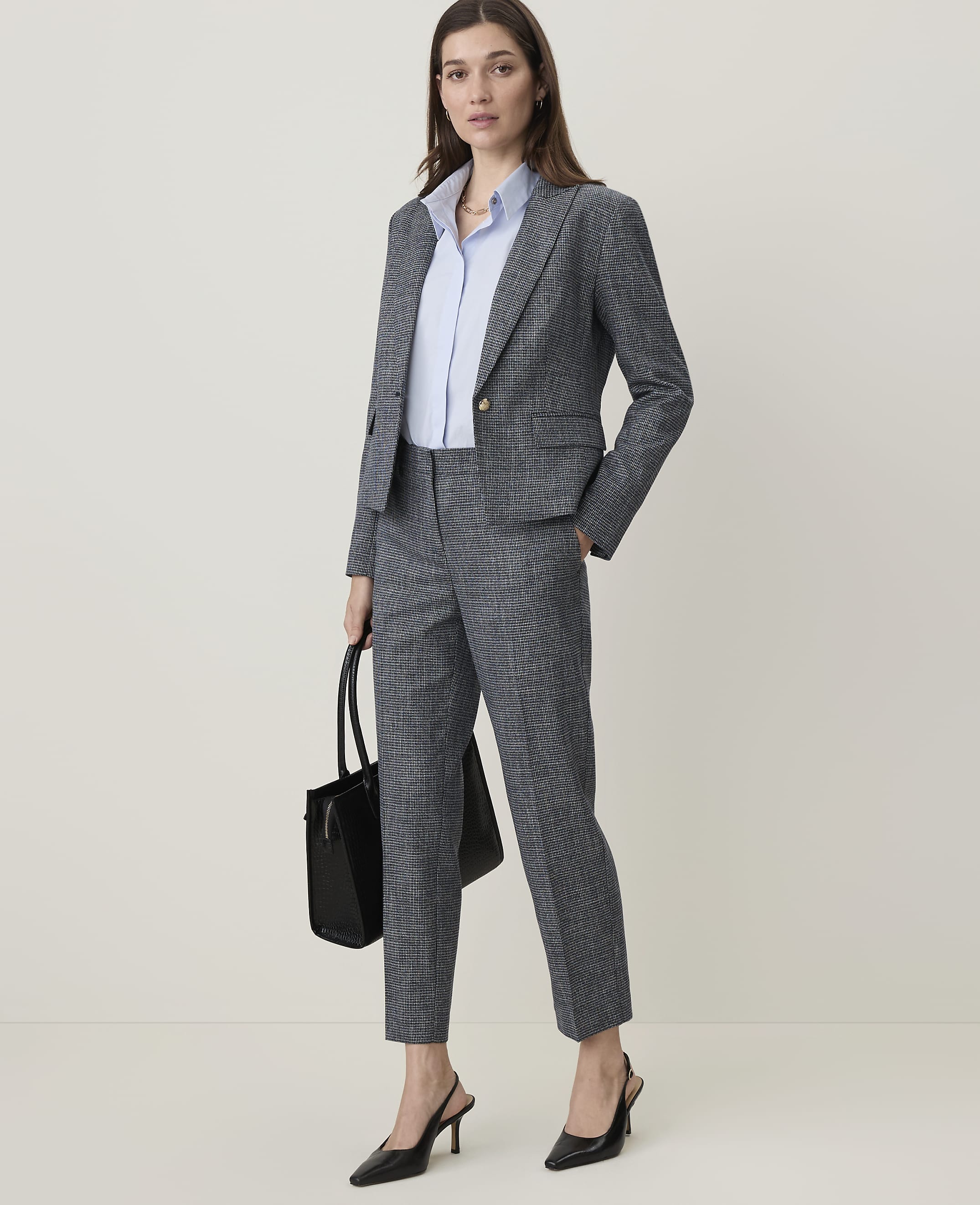 A woman in a grey suit and light blue shirt stands holding a black bag, with a plain, light grey background.