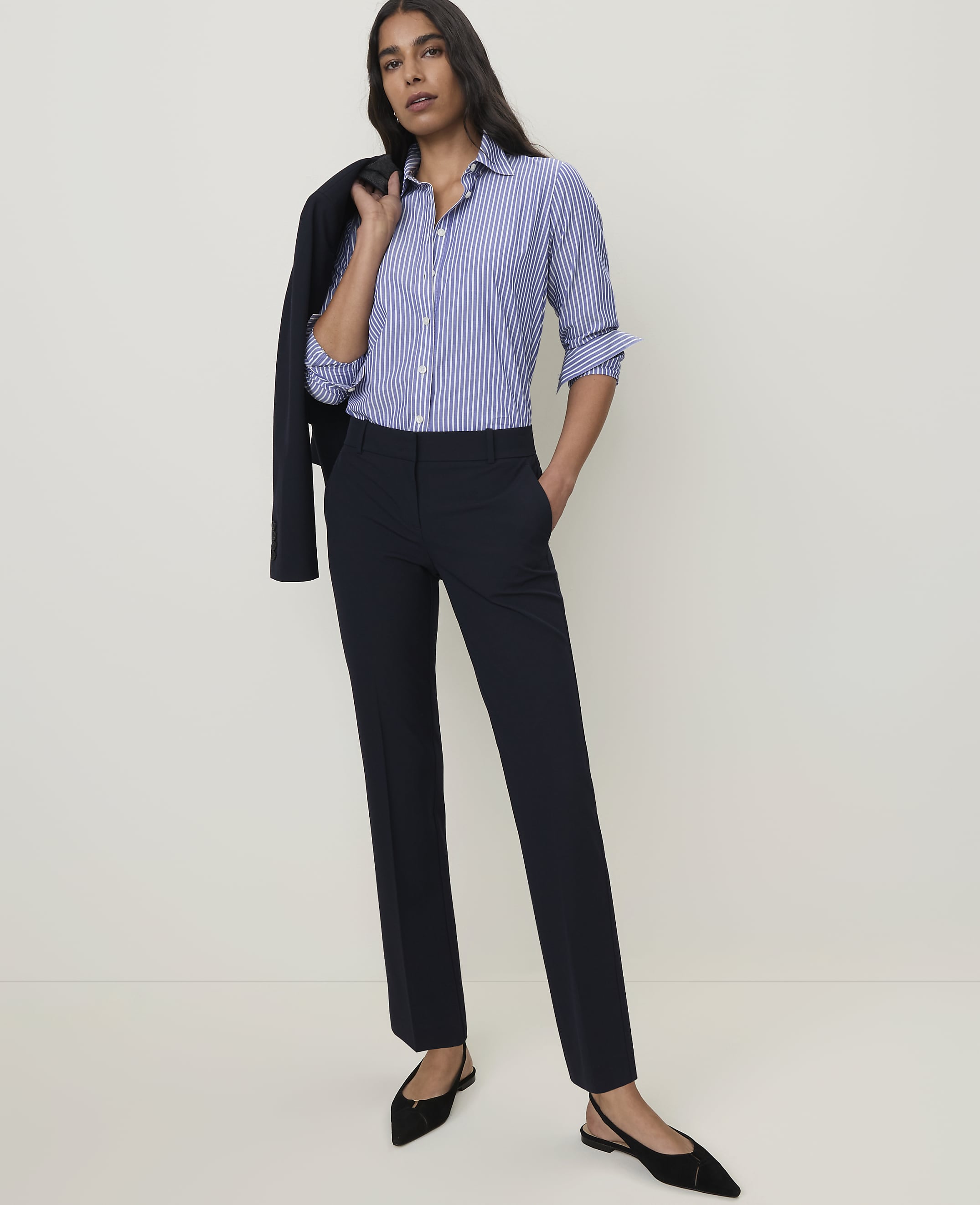 A woman in a striped shirt and dark pants stands against a plain, light background with a dark jacket draped over her shoulder.