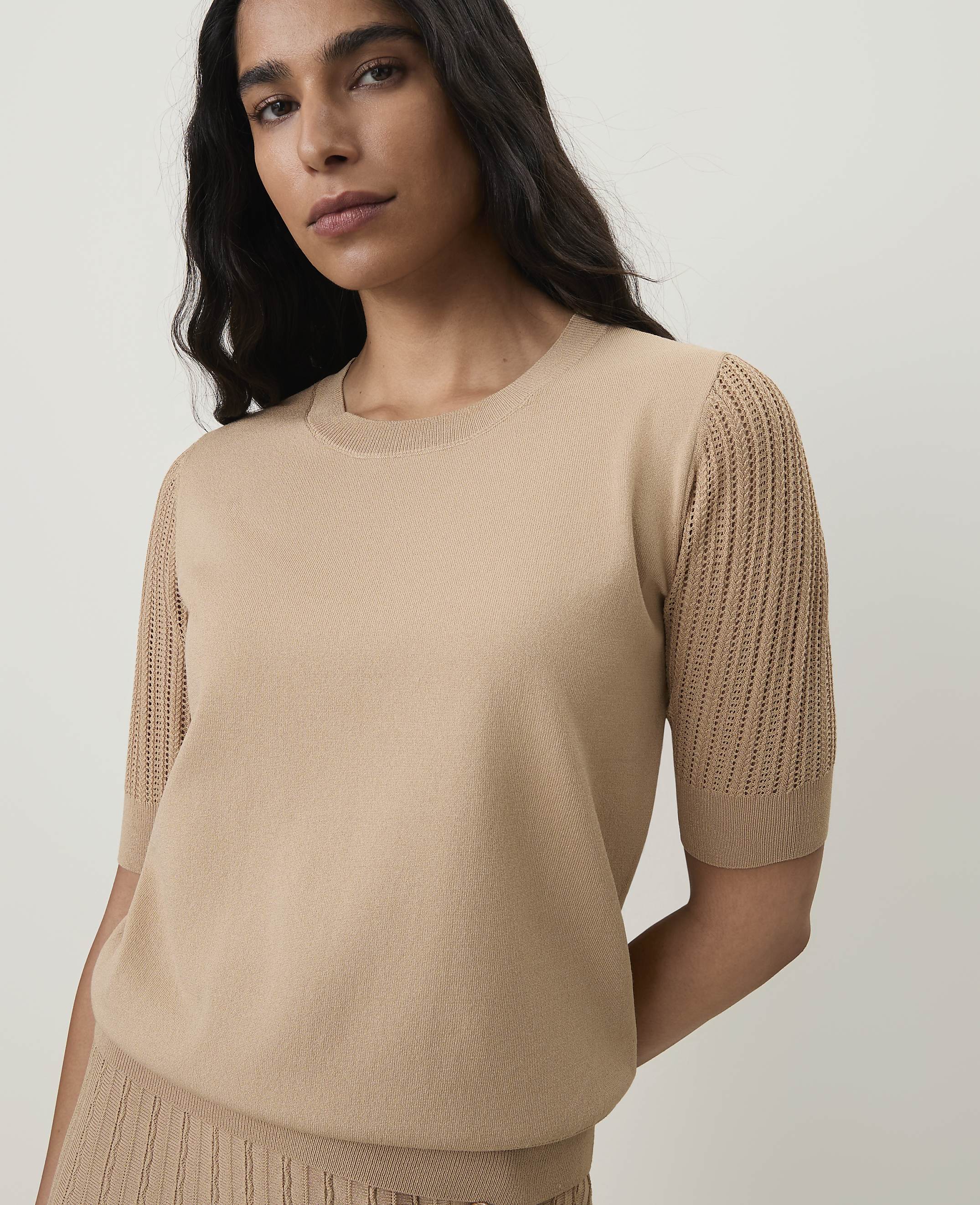 A woman in a beige knit top with openwork sleeves stands against a plain, light background.