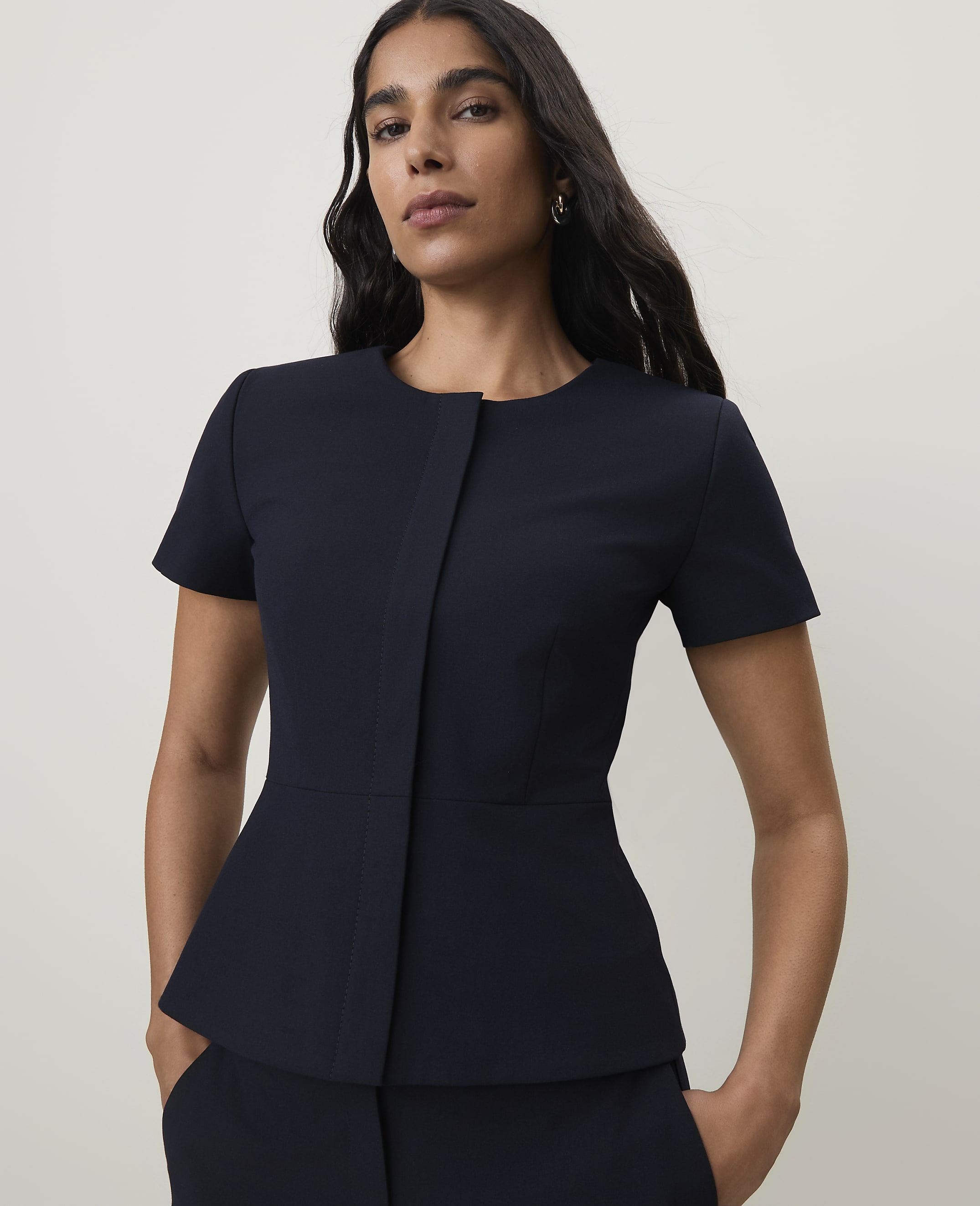 A woman in a navy peplum top and pants stands against a plain, light background.