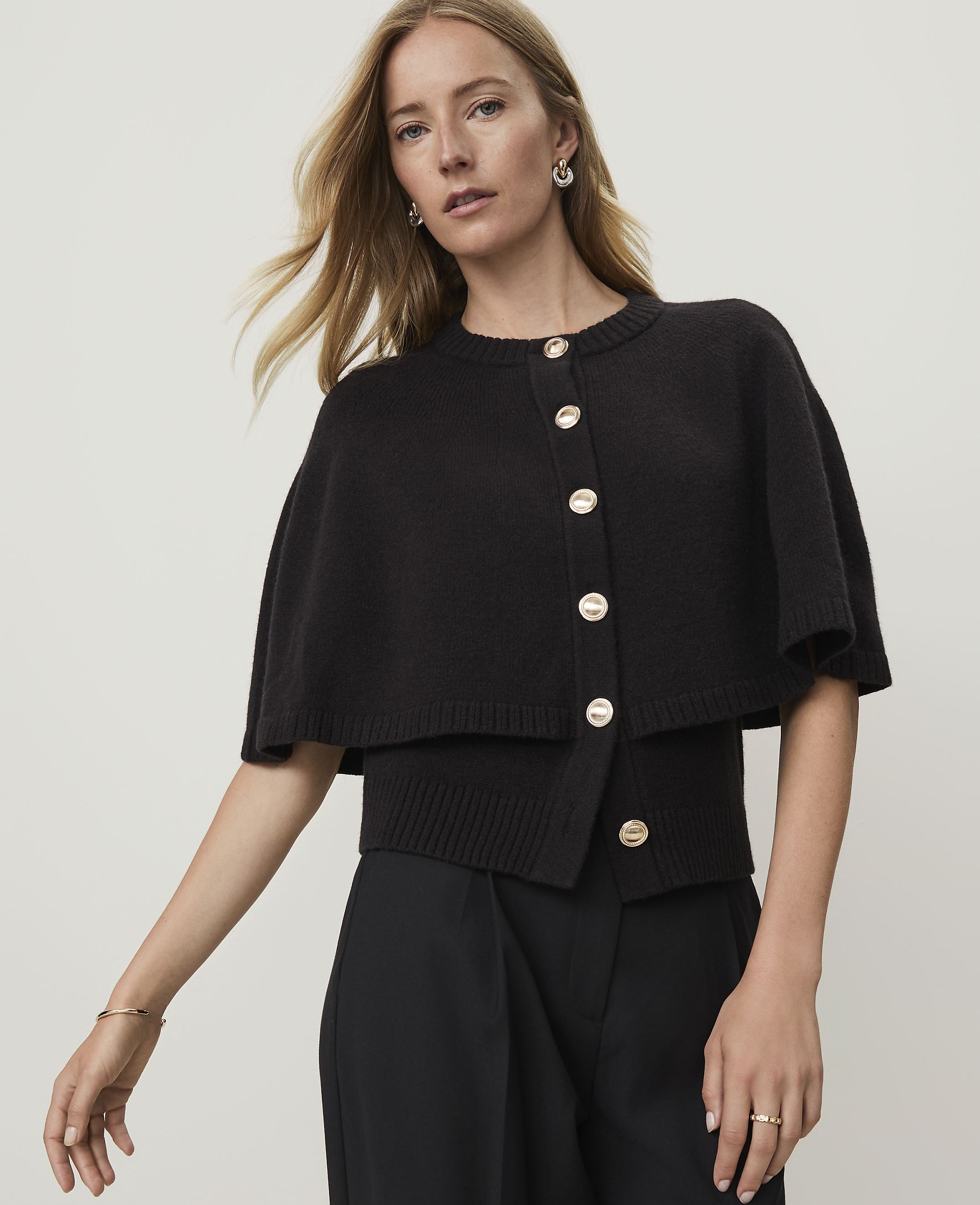 A blonde woman in a black capelet sweater and black pants poses against a plain white background.