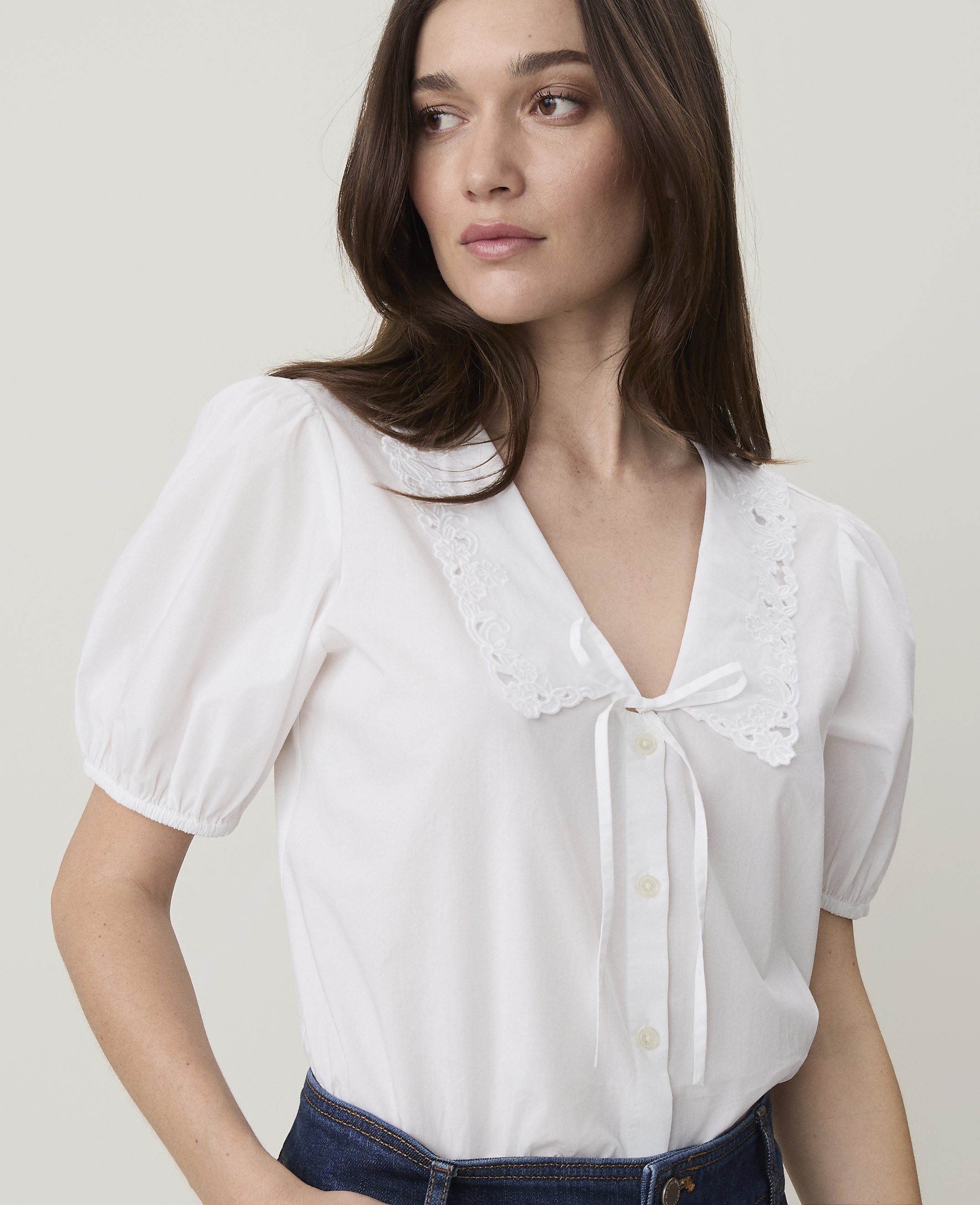 A woman in a white puff-sleeved blouse with a lace collar and a white ribbon tie looks to the side against a plain white background.