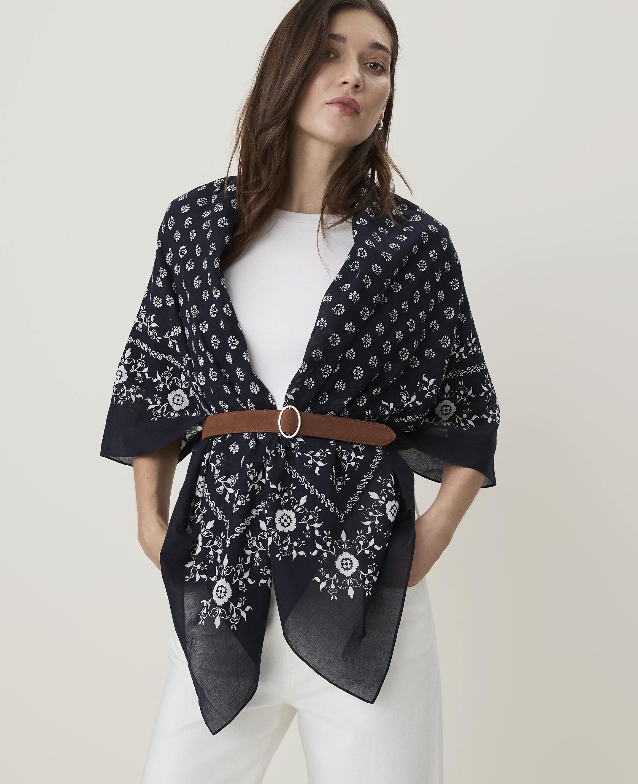 A woman in a white top and white pants wears a dark blue patterned scarf cinched with a brown belt, against a plain white background.
