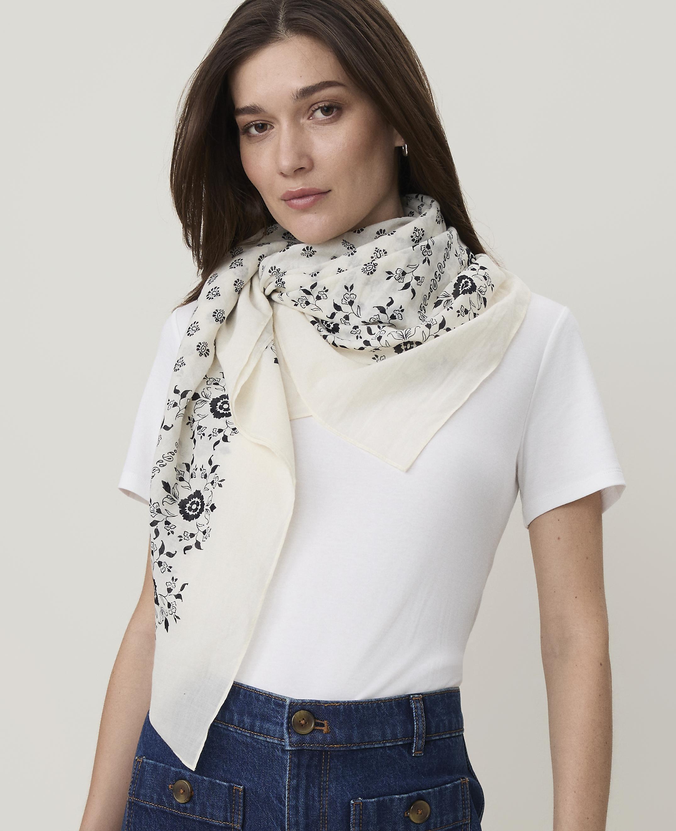 A woman in a white t-shirt and denim skirt models a cream-colored scarf with a black floral pattern against a plain white background.