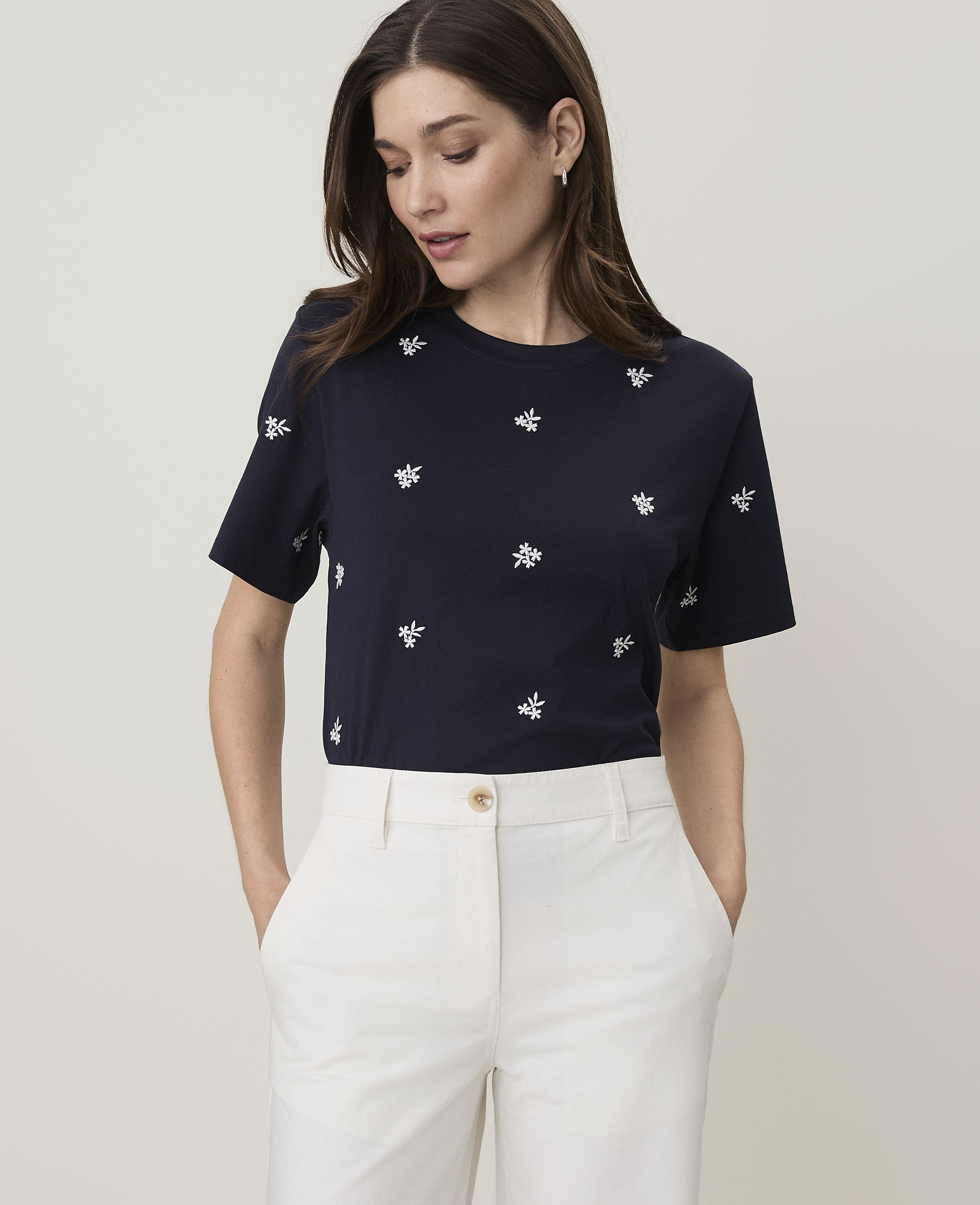 A woman in a navy t-shirt with embroidered bees and white pants stands against a plain, light gray background.