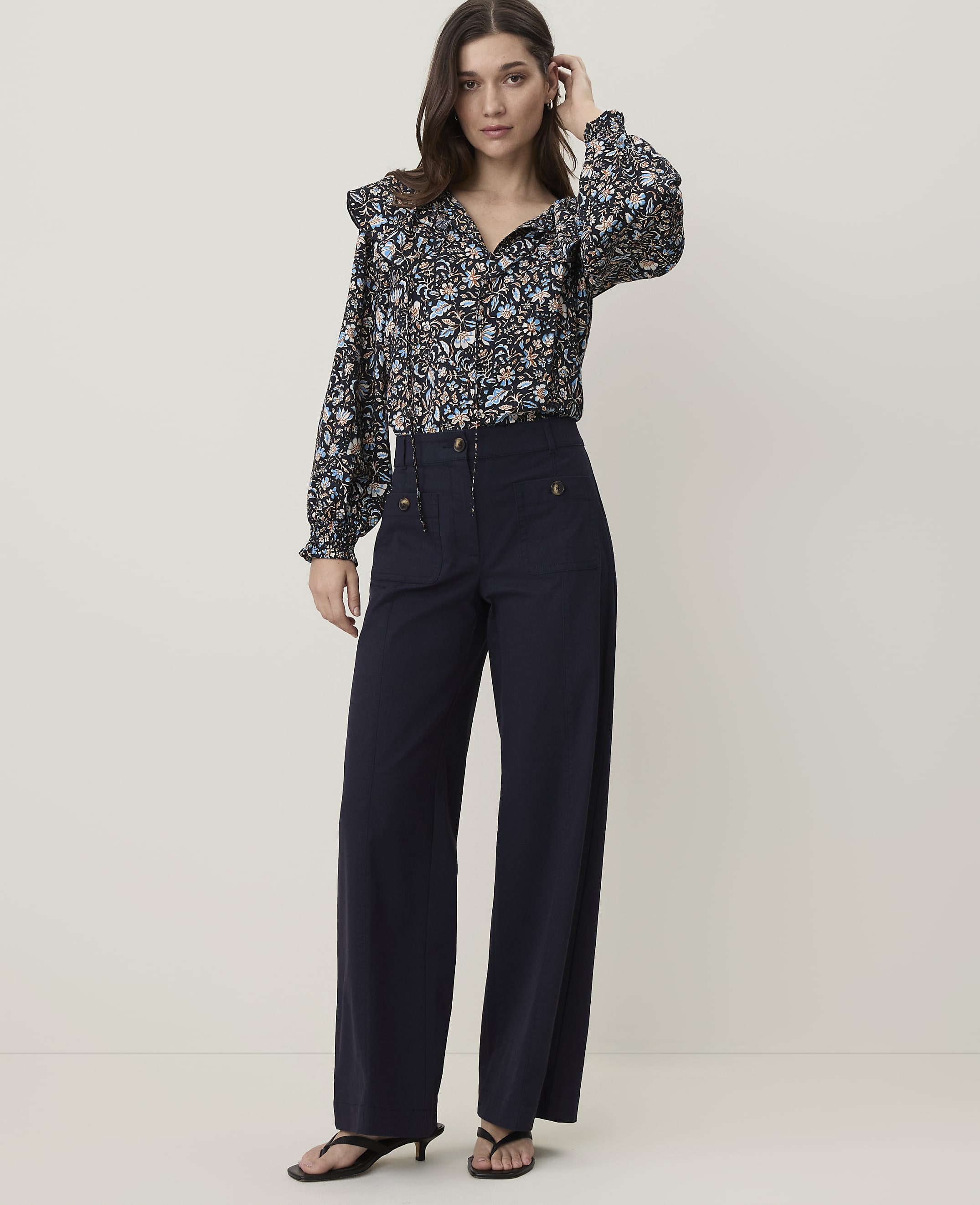 A woman in a floral blouse and navy wide-leg pants stands against a plain, light-colored background.