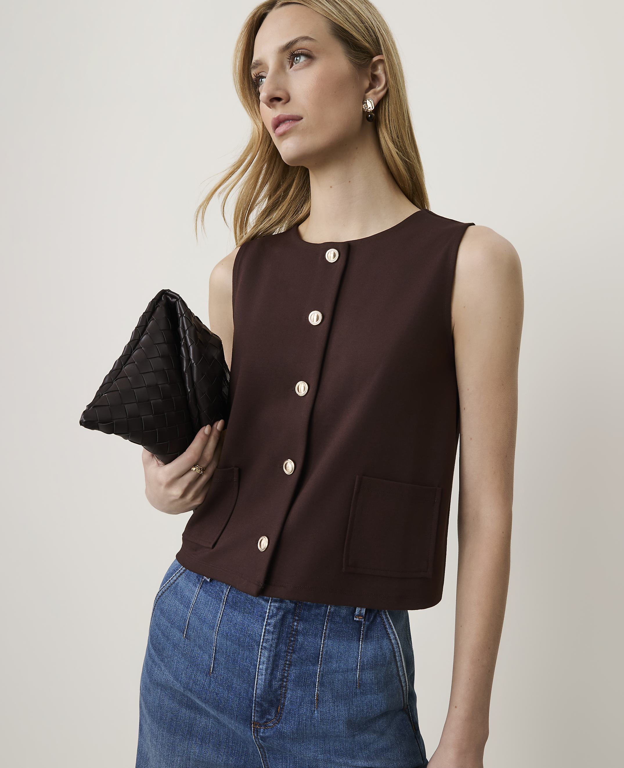 A woman in a brown sleeveless top and blue jeans holds a black clutch in front of a plain background.