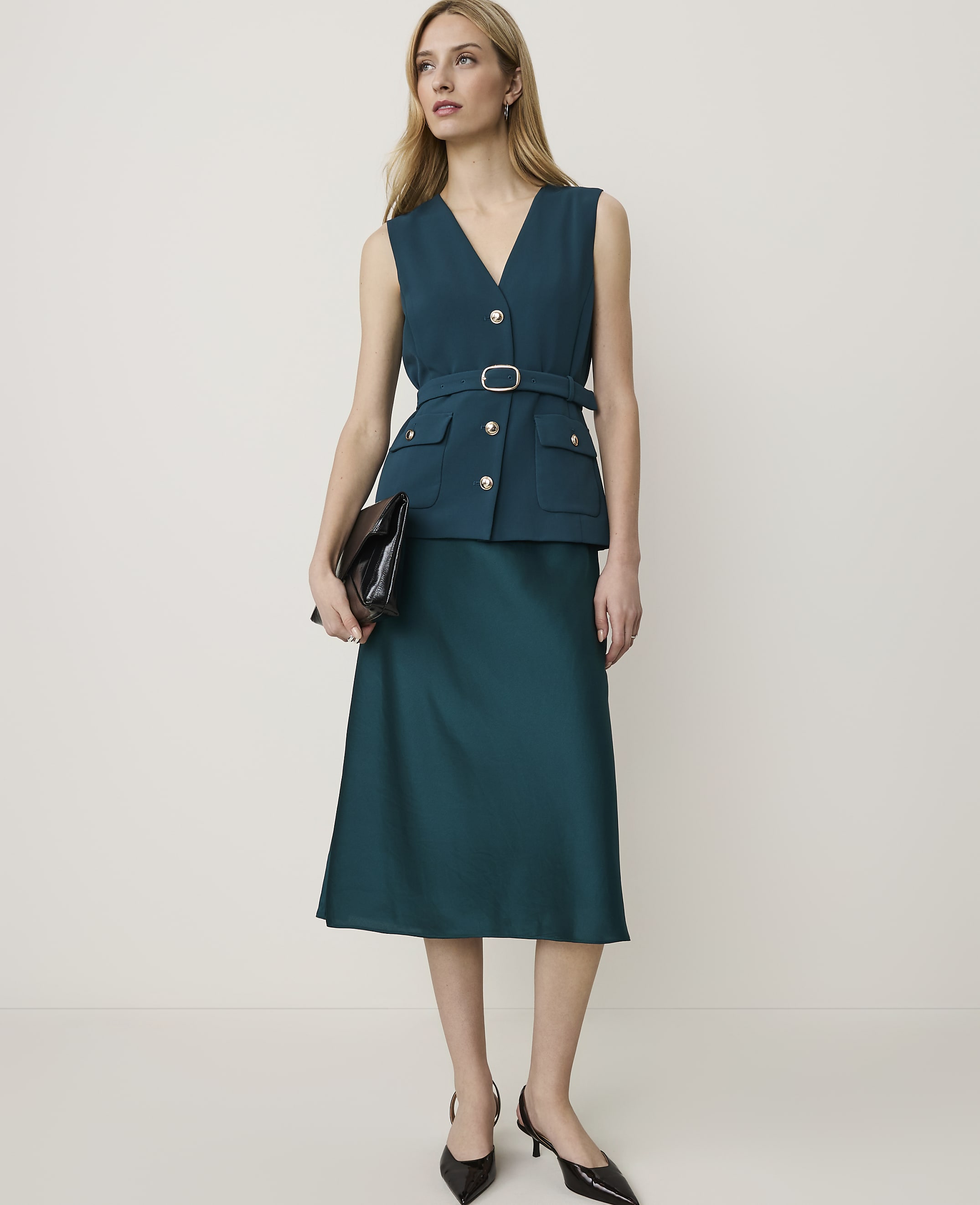 A woman in a teal vest and skirt stands against a plain white background, holding a dark clutch.