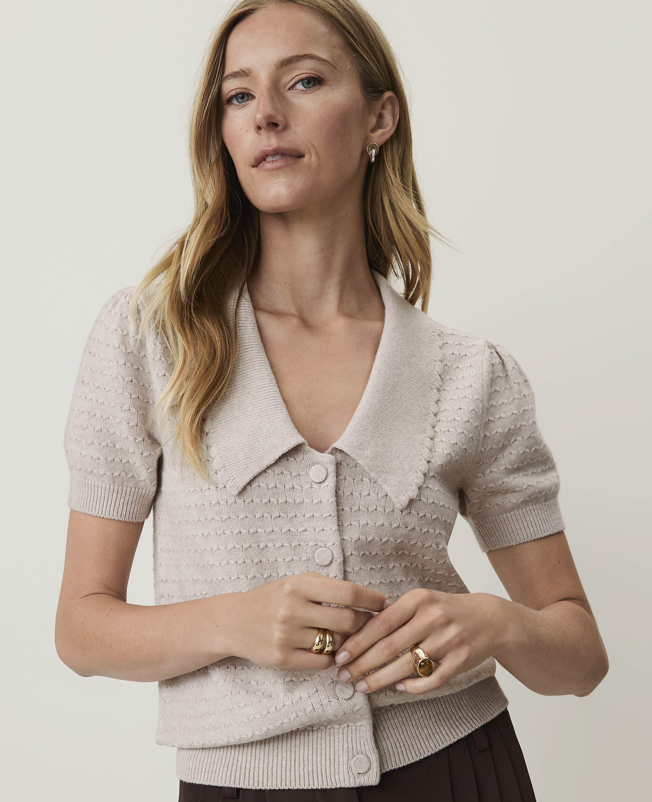 A woman in a beige cardigan with a Peter Pan collar stands against a plain, light background.