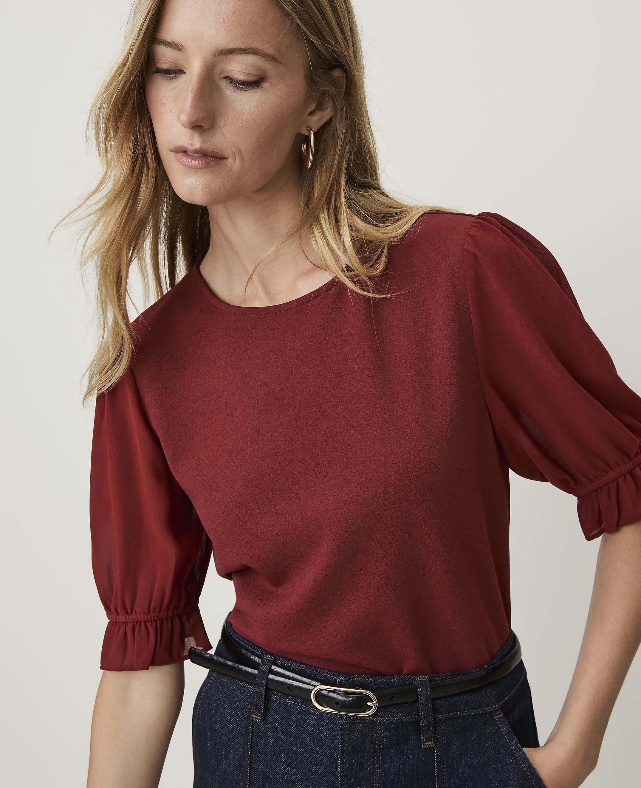A woman in a rust-colored top with ruffled sleeves and a black belt stands against a plain white background.