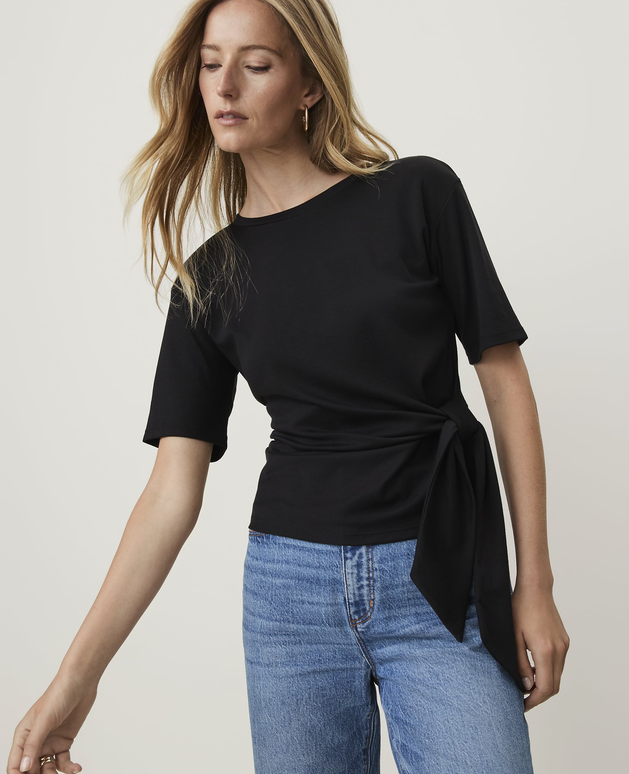 A blonde woman in a black tie-waist top and blue jeans poses against a plain gray background.