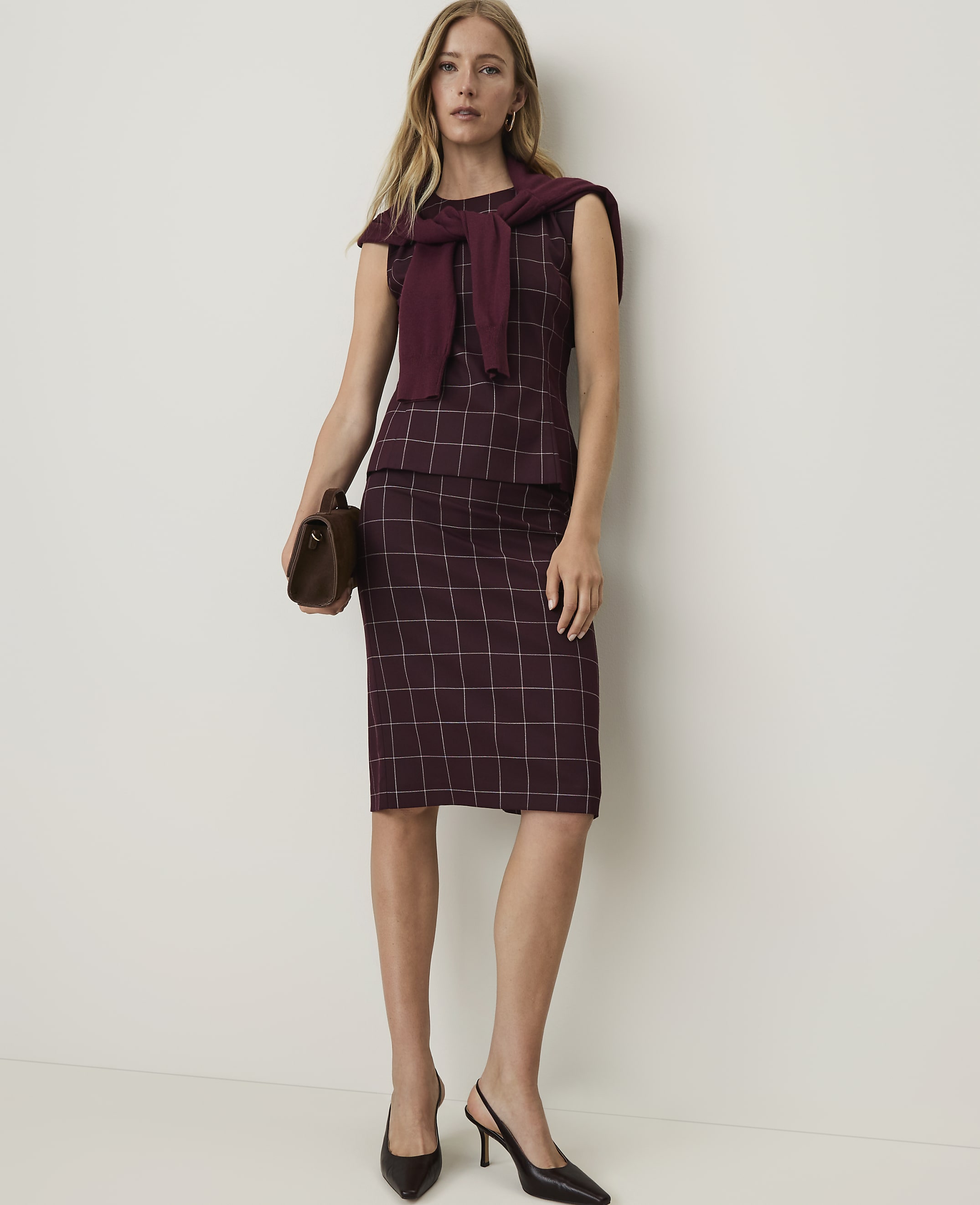 A woman in a burgundy plaid dress and sweater draped over her shoulders stands against a plain white background, holding a brown clutch.