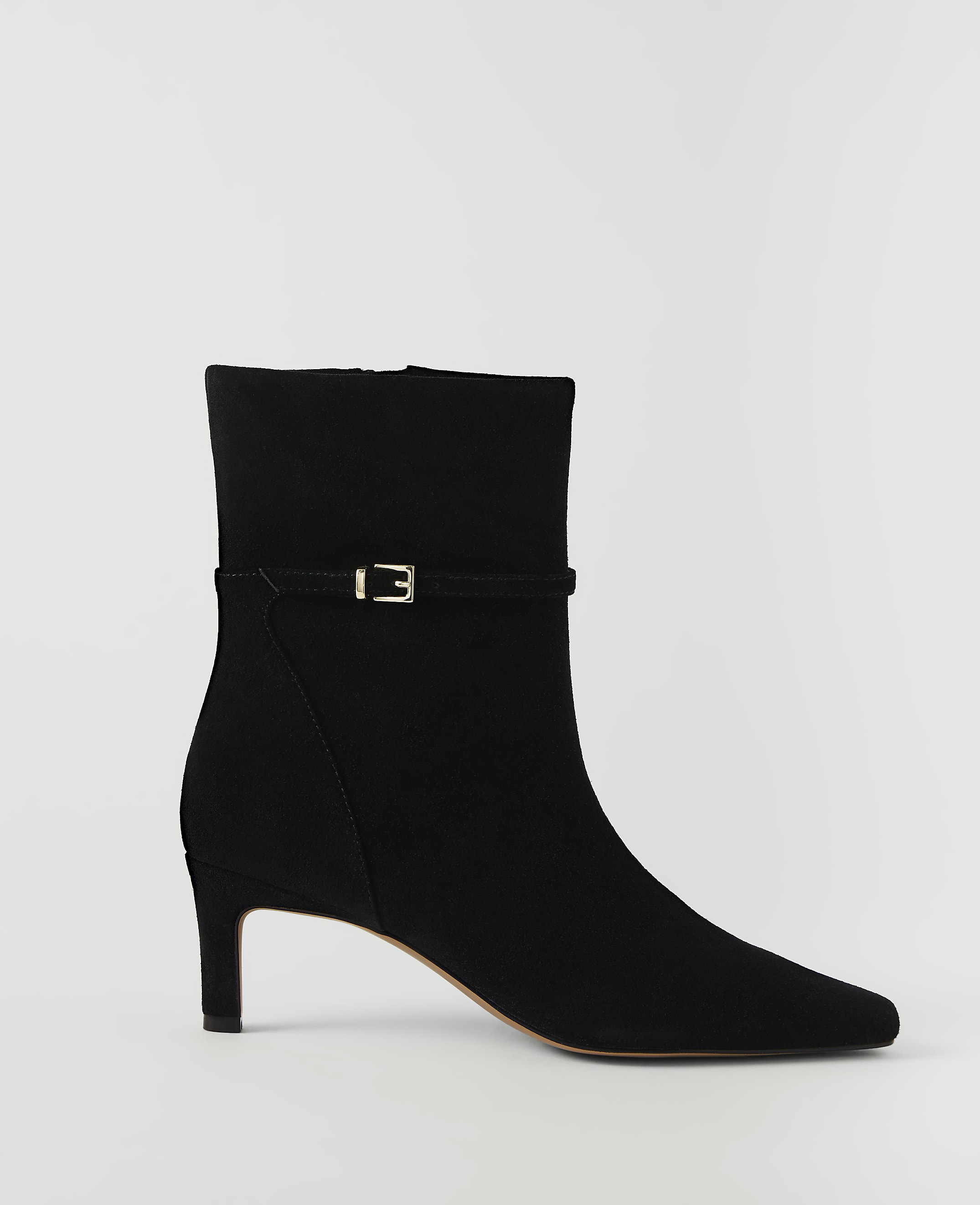 A black suede ankle boot with a gold buckle detail stands against a plain white background.