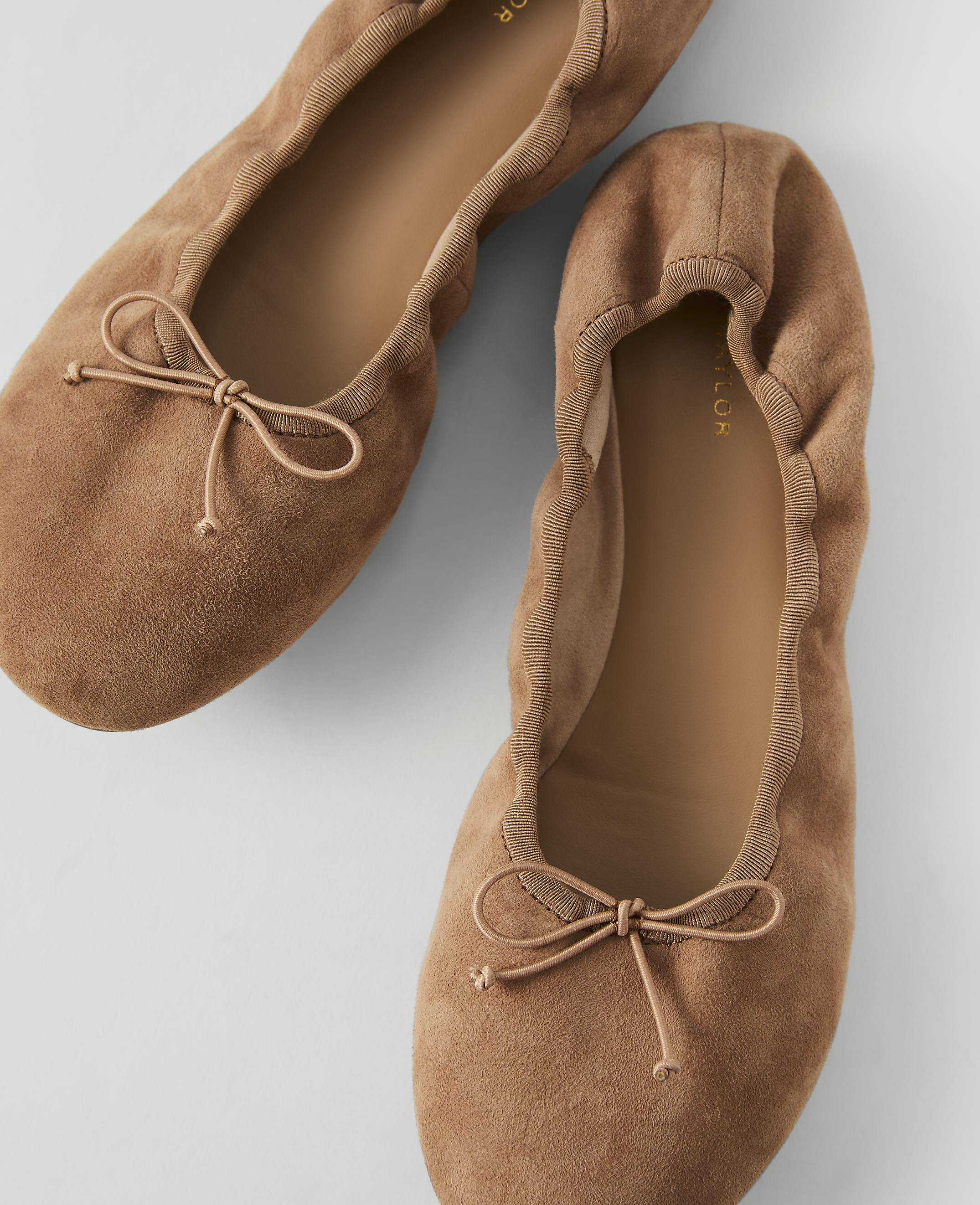 Two tan suede ballet flats with bows are arranged on a light gray background.