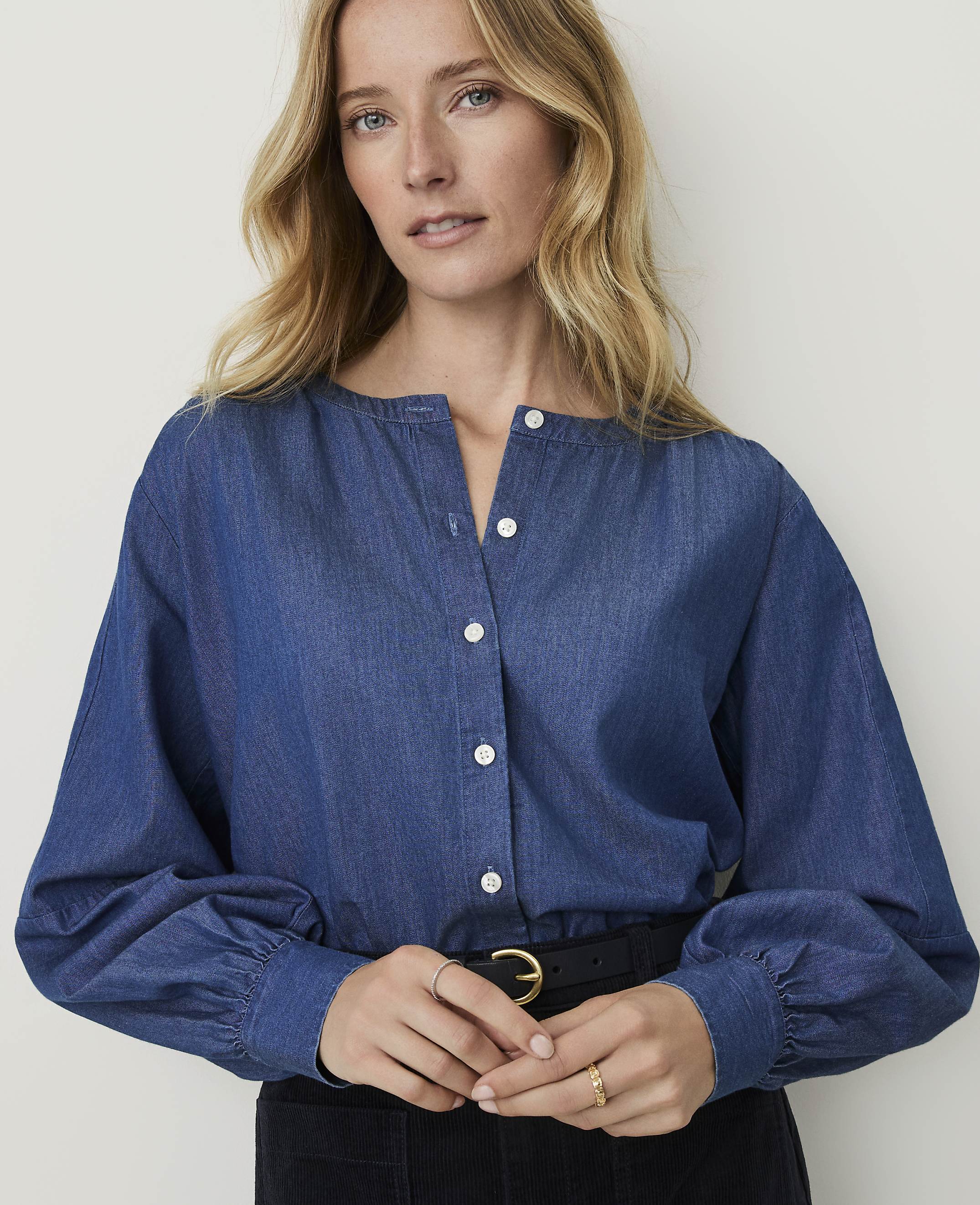 A blonde woman in a blue denim shirt with puffed sleeves stands against a plain, light-colored background.