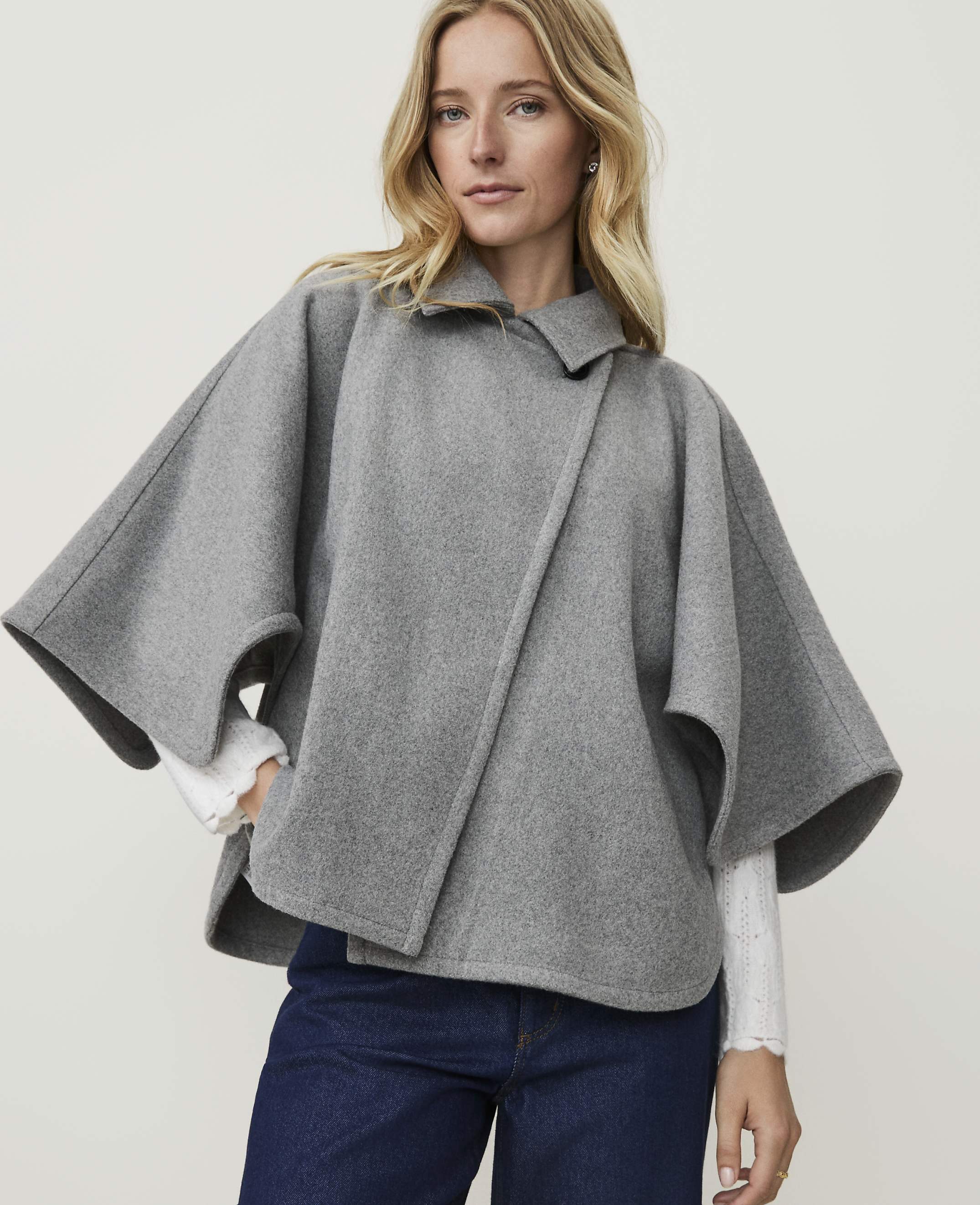 A blonde woman in a grey cape and blue jeans stands against a plain white background.