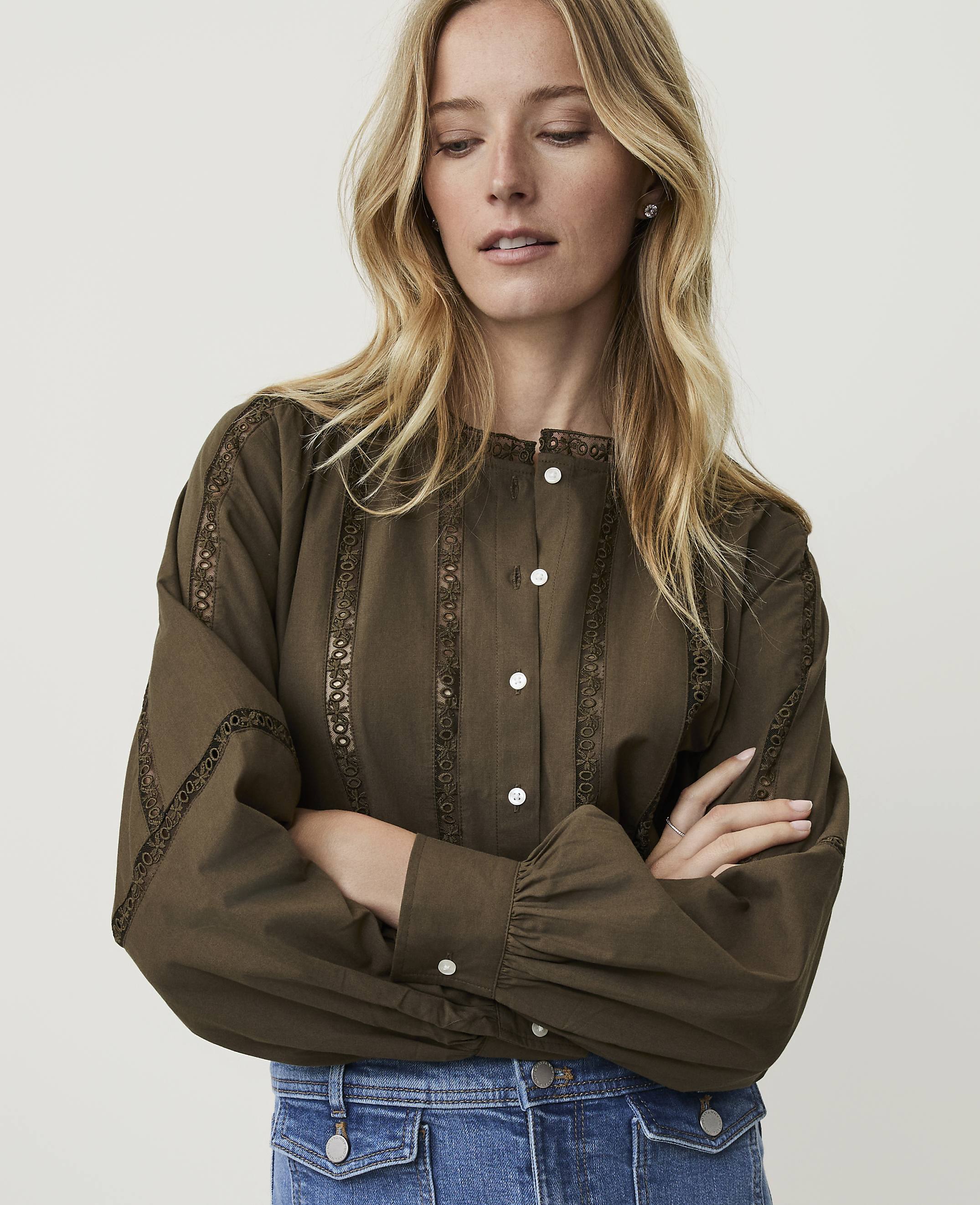 A blonde woman in a green blouse and blue jeans stands with her arms crossed against a plain white background.