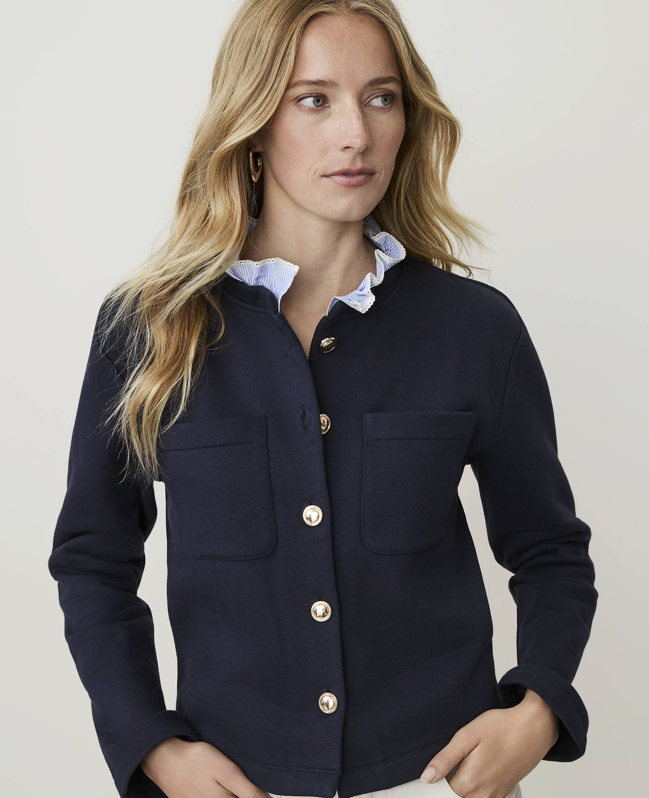 A blonde woman in a navy blue jacket with gold buttons and a ruffled collar stands against a plain white background.
