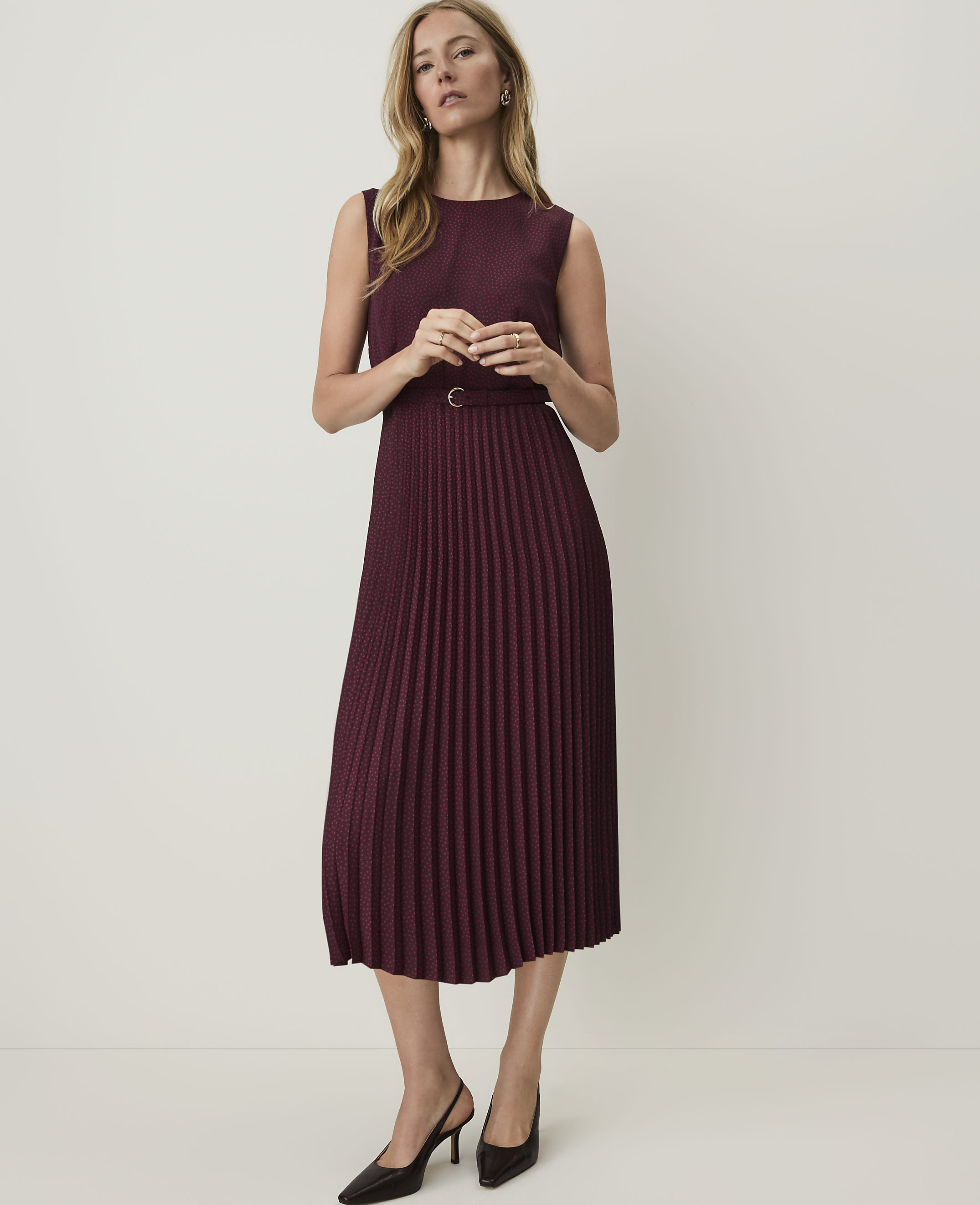 A woman in a burgundy pleated dress and black heels stands against a plain, light background.