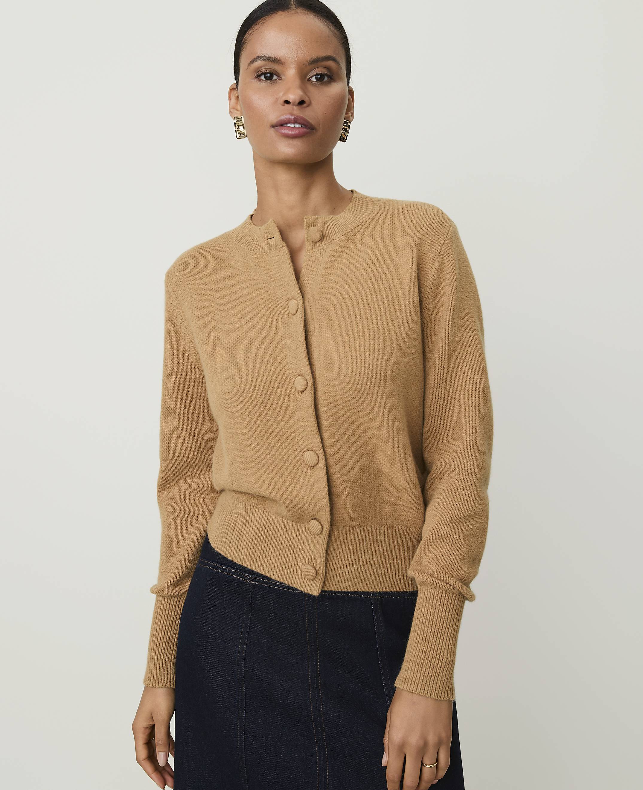 A woman in a tan cardigan and dark skirt stands against a plain background.