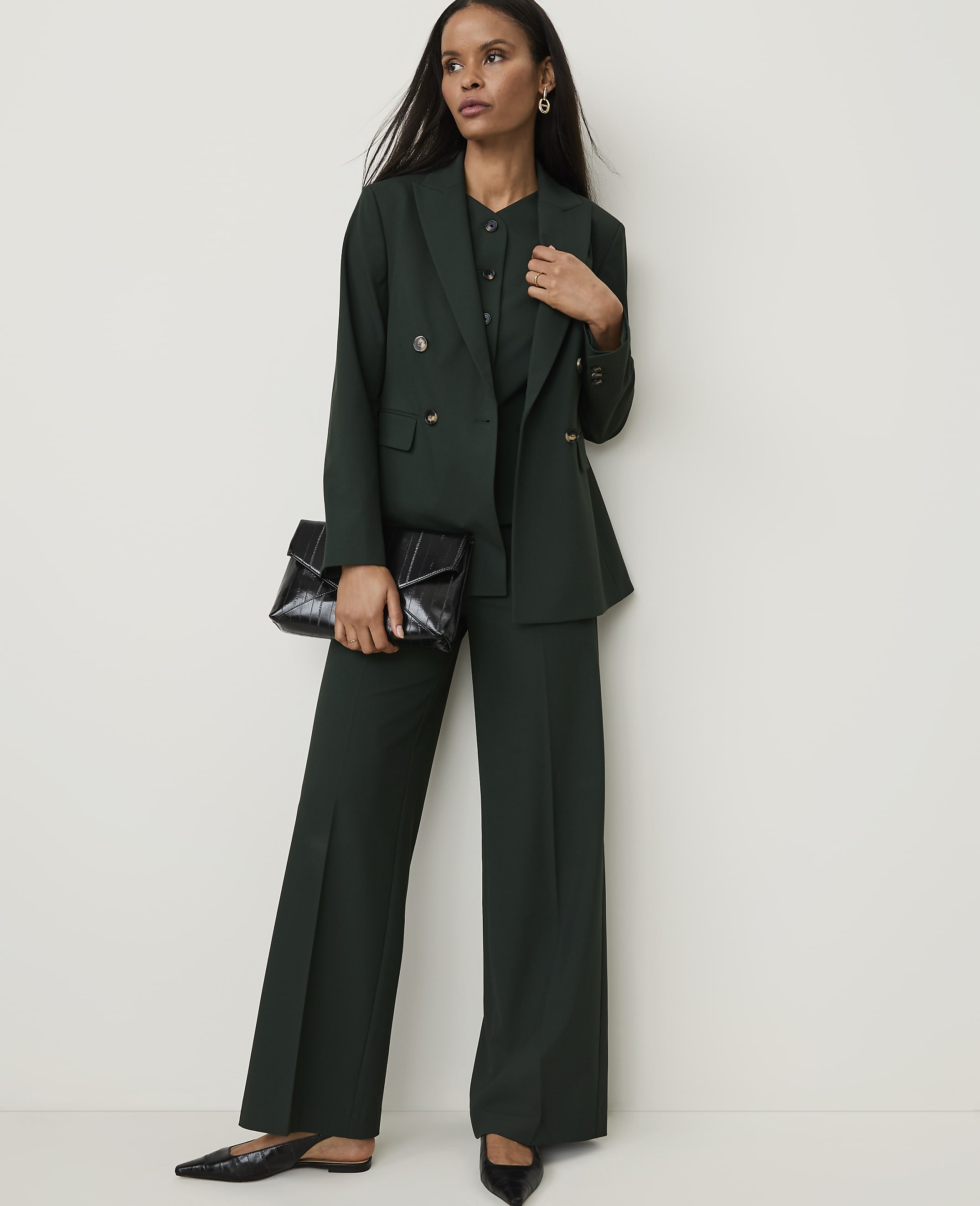 A woman in a dark green pantsuit and holding a black clutch stands against a plain white background.