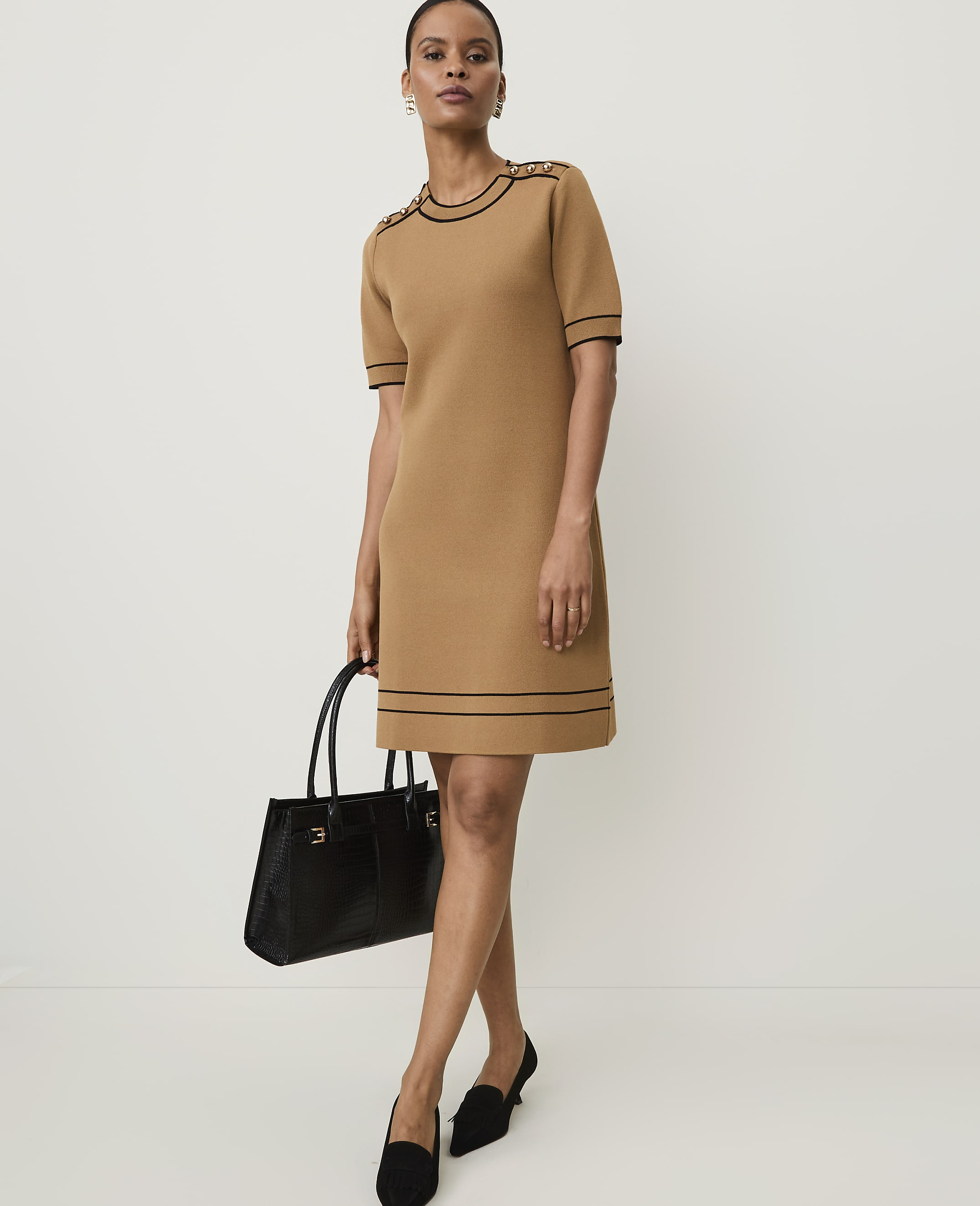 A woman in a tan knit dress with black trim stands holding a black handbag against a plain white background.