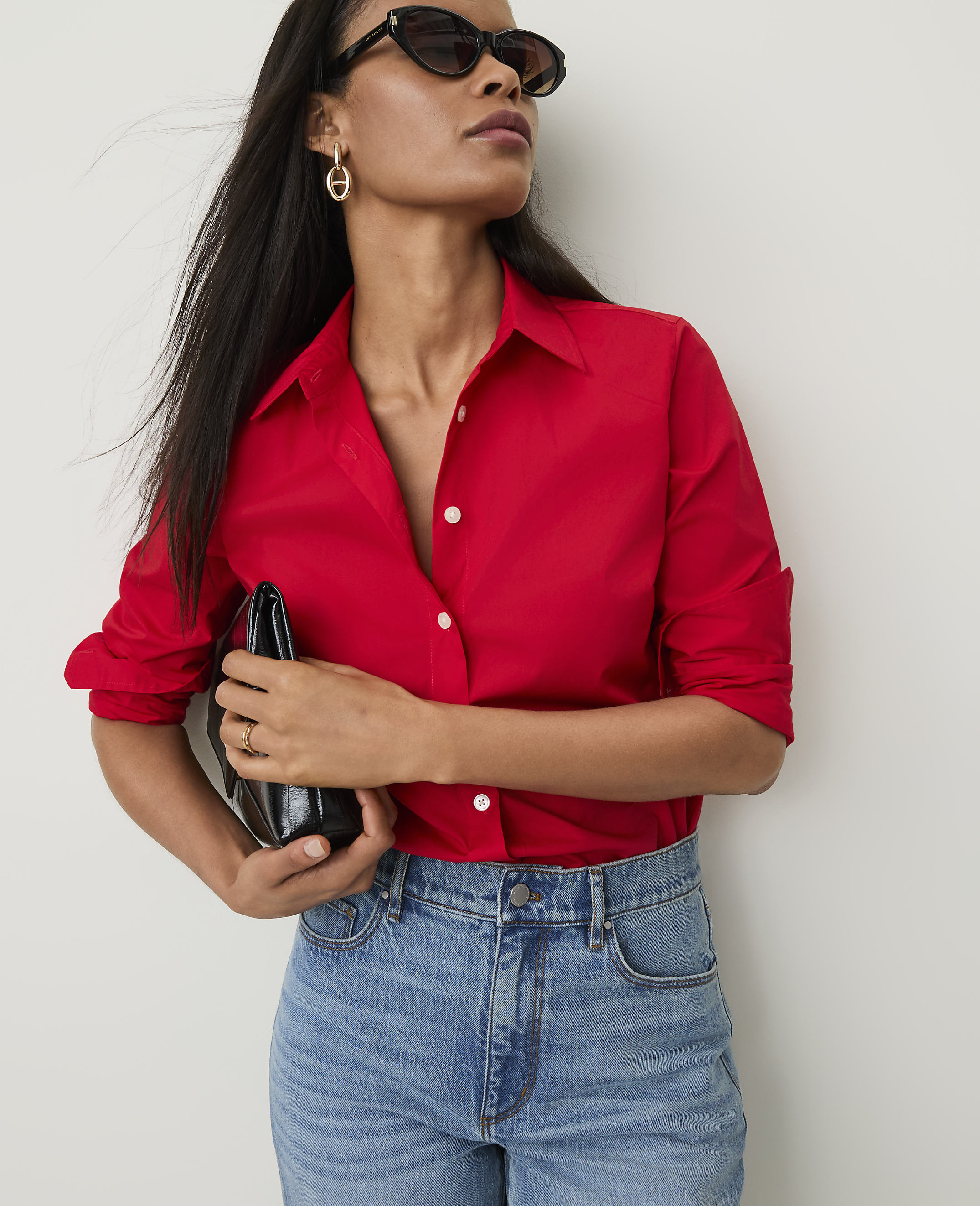 A woman in a red shirt and jeans holds a black clutch against a plain white background.