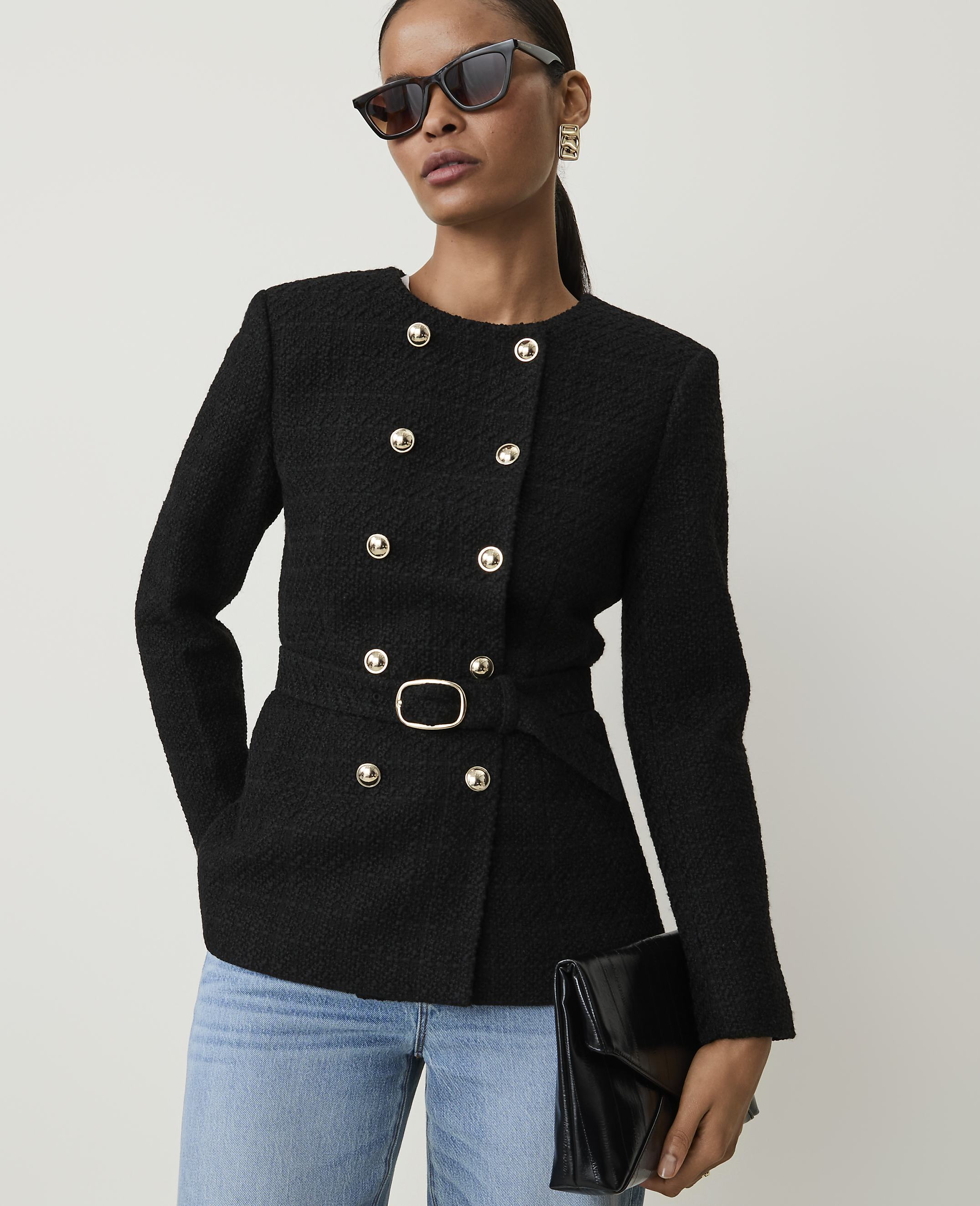 A woman in a black tweed jacket and jeans holds a black clutch against a plain white background.