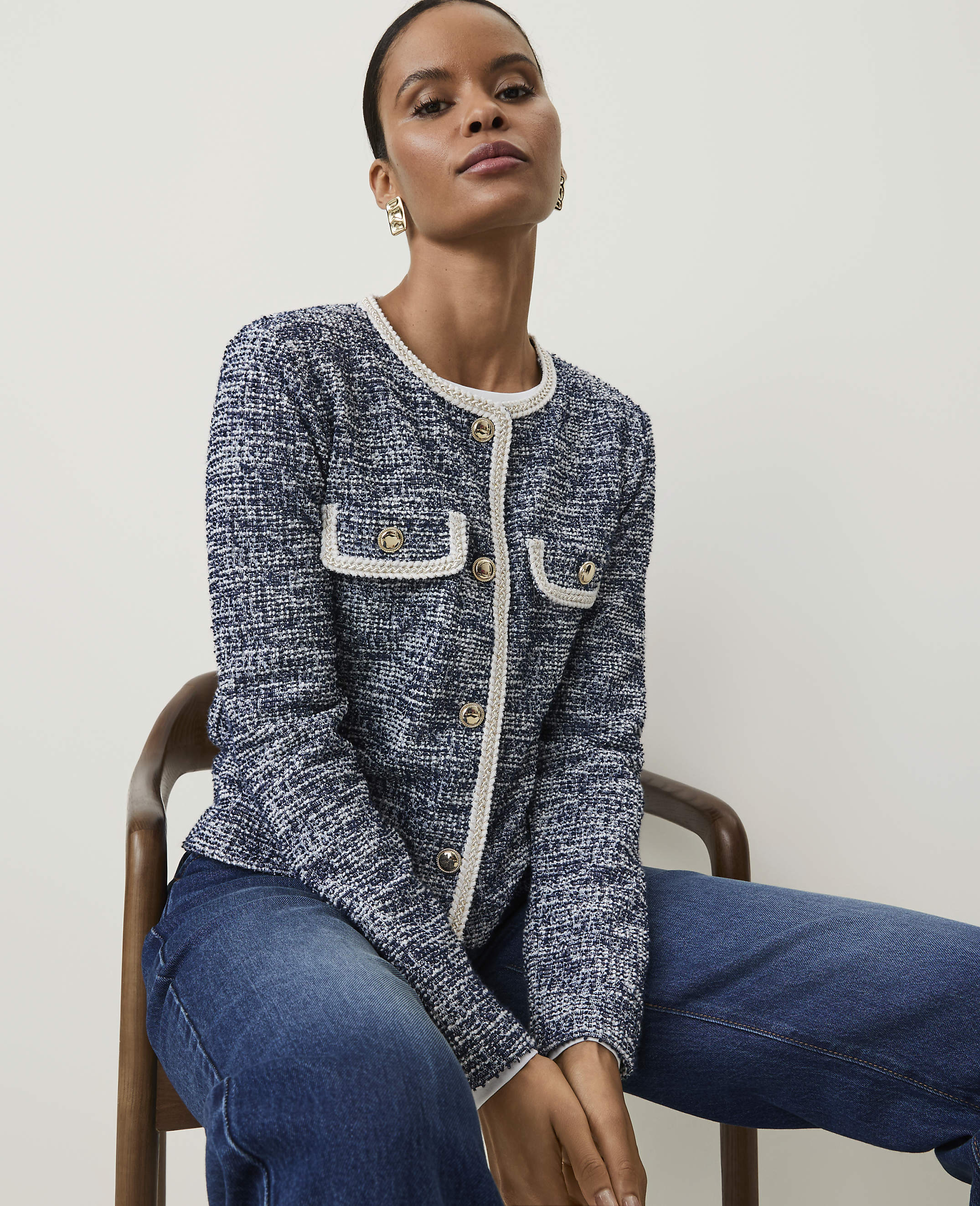 A woman in a blue tweed jacket and jeans sits on a wooden chair against a plain white background.