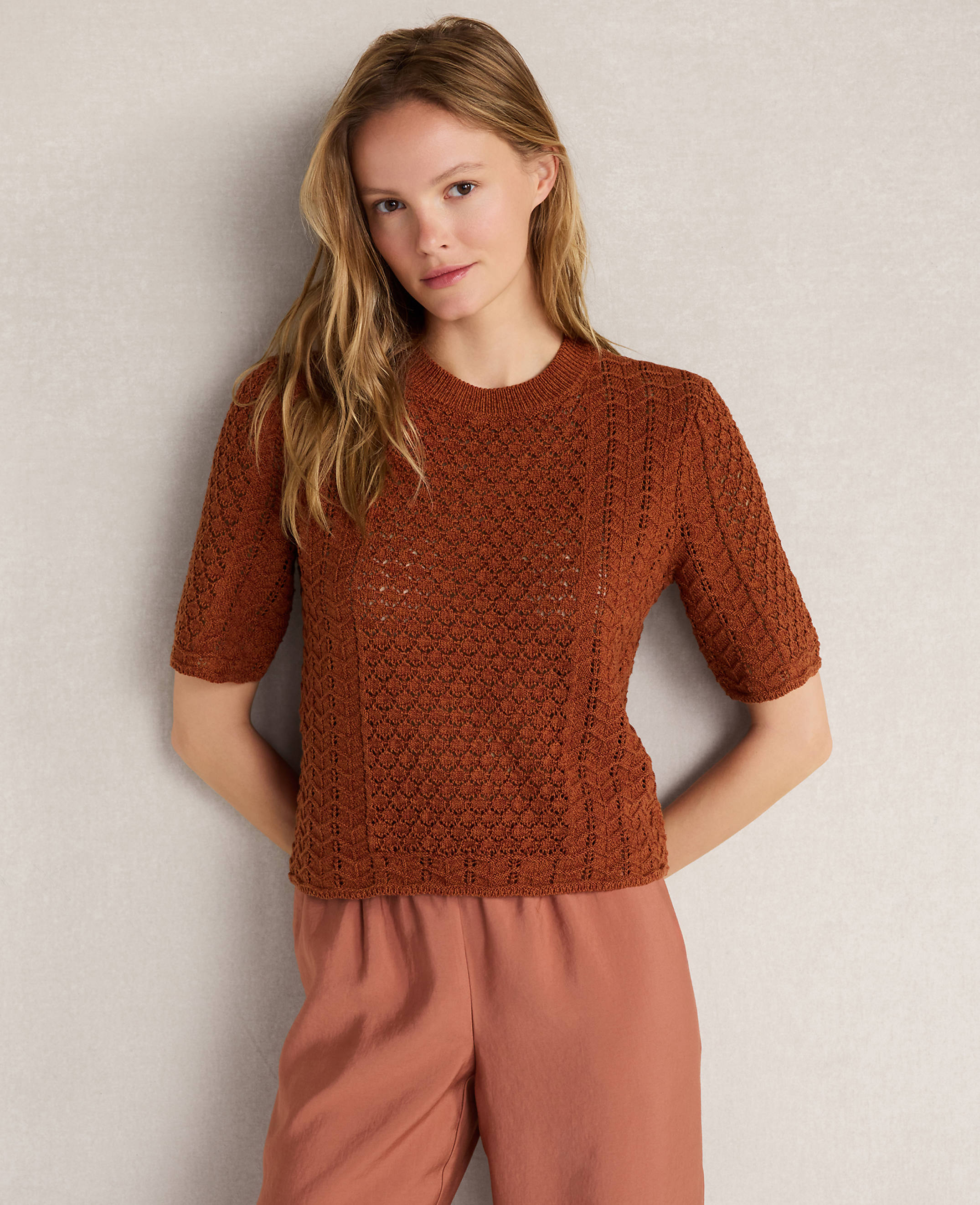 A young woman in a brown knit top and pink pants stands against a plain gray background.